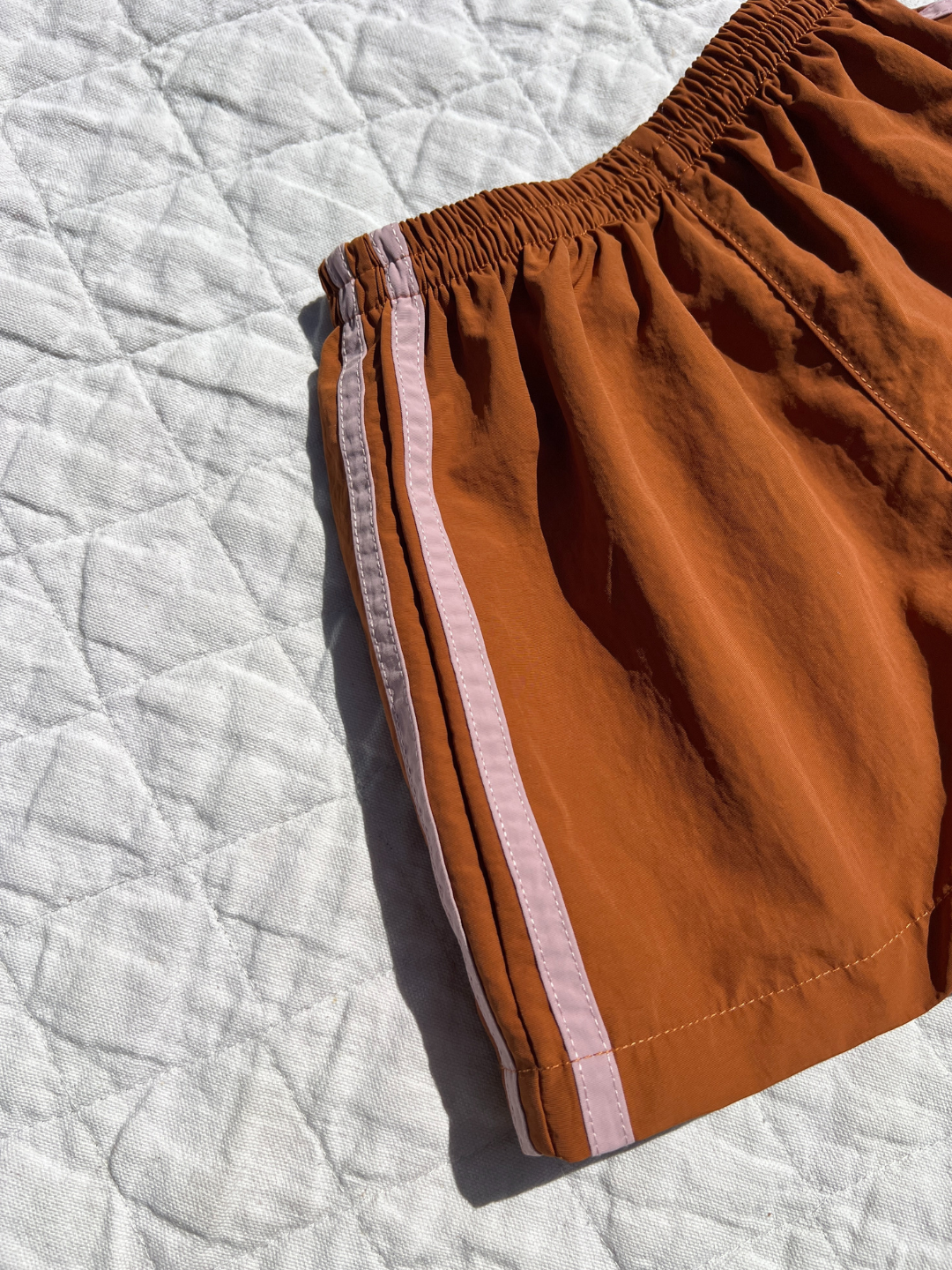Rust | The SPRINTER SHORTS, featuring a vibrant orange color, light pink side stripe, and elasticated waist, are displayed on a textured white quilt in bright sunlight.