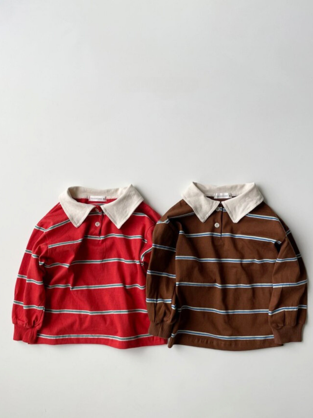 Two RETRO RUGBY LONGSLEEVE tees from a Korean kids brand—one red with white stripes, the other brown with blue and white stripes, both featuring classic rugby collars—are laid out side by side.