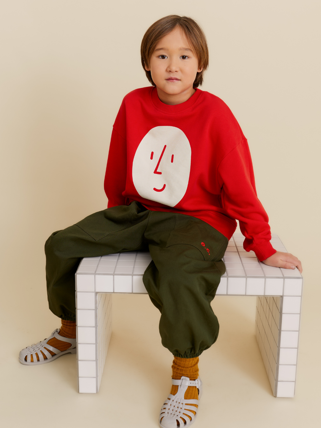 Red | A child with straight brown hair sits on a white tiled bench, wearing a red POTATO SWEATSHIRT with a face graphic, olive green pants, orange socks, and white sandals.