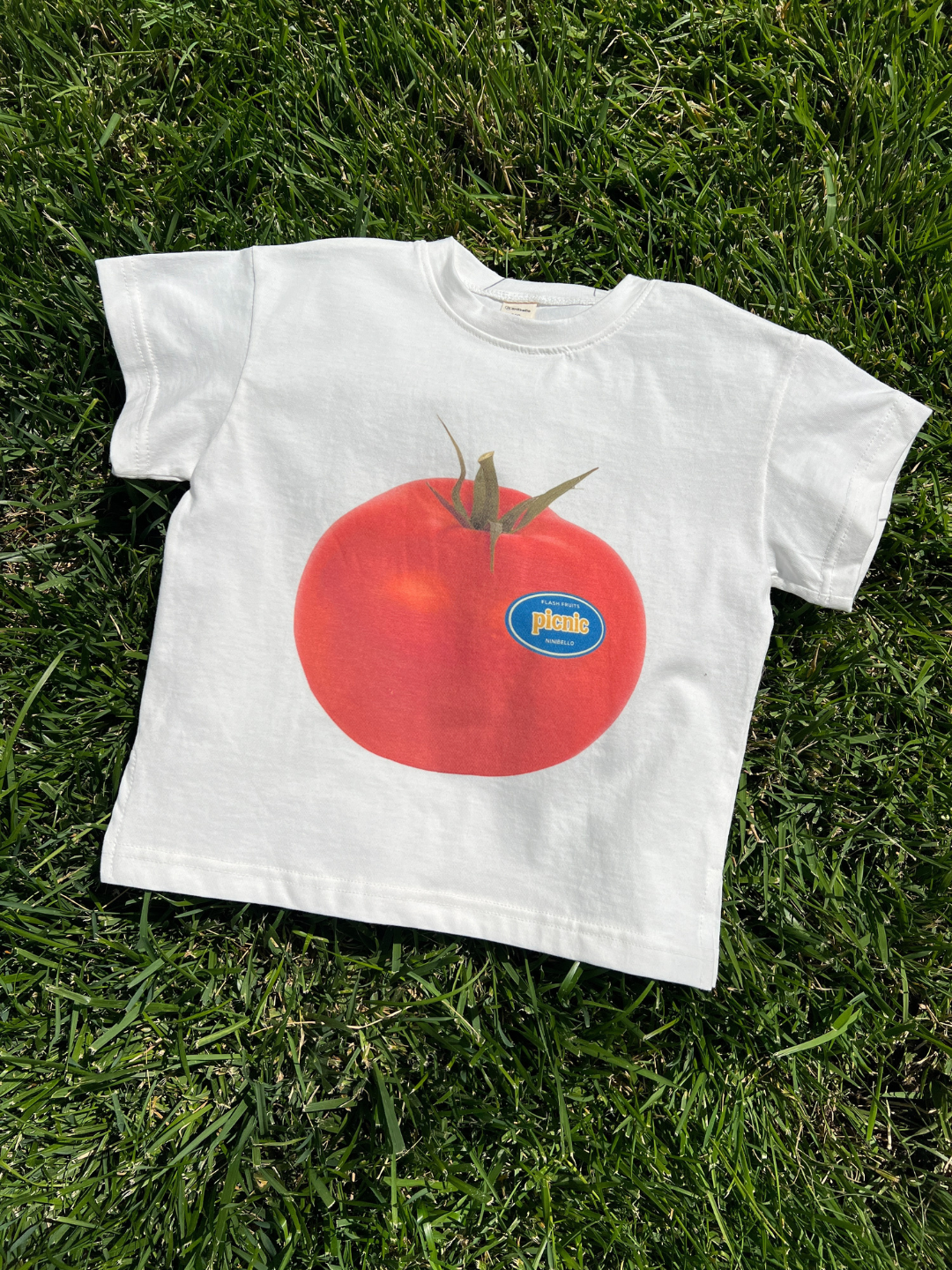 The PICNIC TEE is an oversized cotton t-shirt from a Korean kids brand, featuring a bold screen-printed tomato graphic and a small blue-and-yellow "prime" sticker, laid flat on green grass.