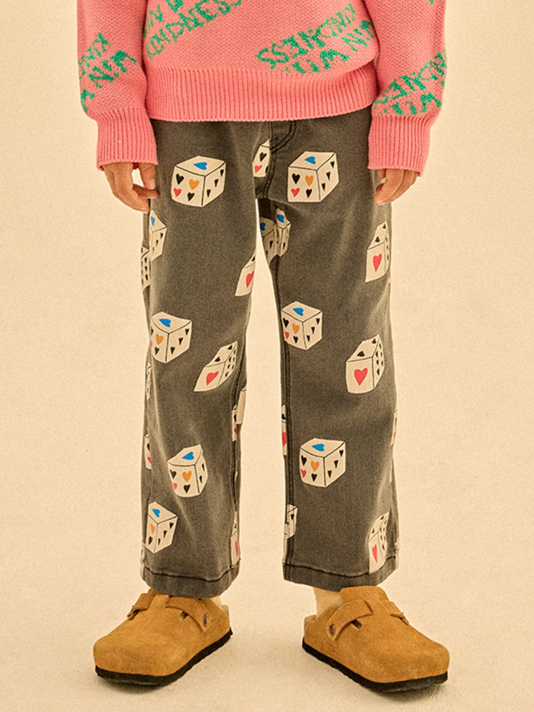 Wearing HEART DICE DENIM PANTS, a pink sweater with green text, beige socks, and tan suede clogs, this person stands against a neutral background.
