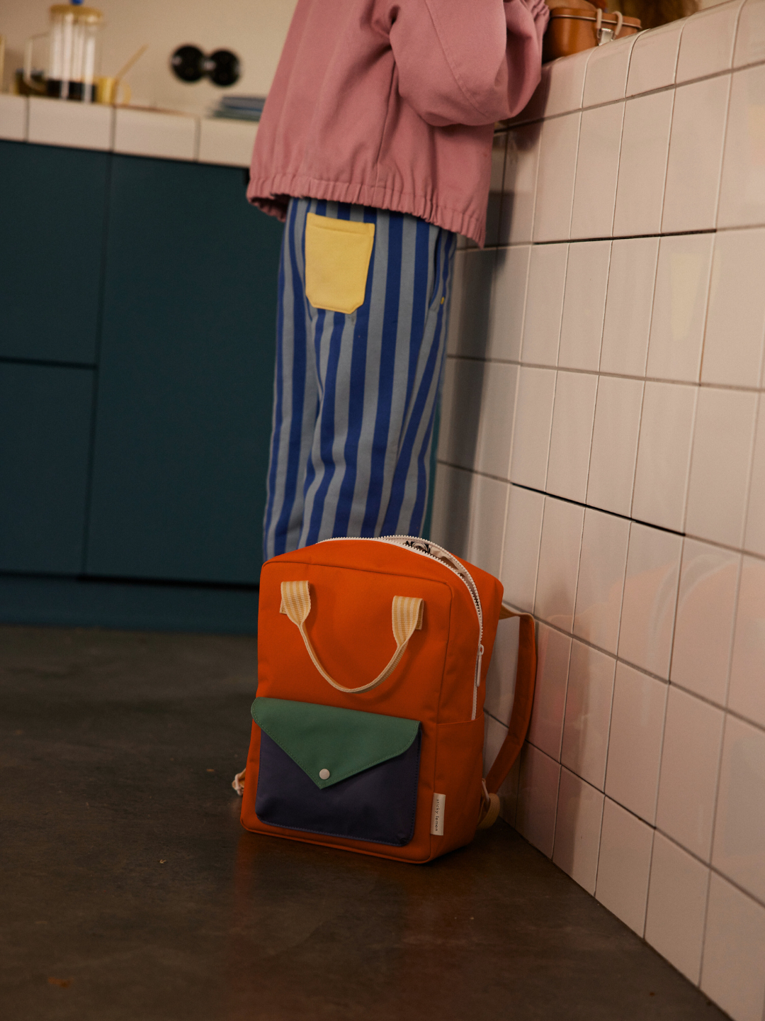 Lifebuoy Orange | A person by a tiled counter wears blue striped pants with a yellow pocket; beside them on the floor is the MEDIUM ENVELOPE BACKPACK, made from recycled materials with colorful pockets and waterproof lining.