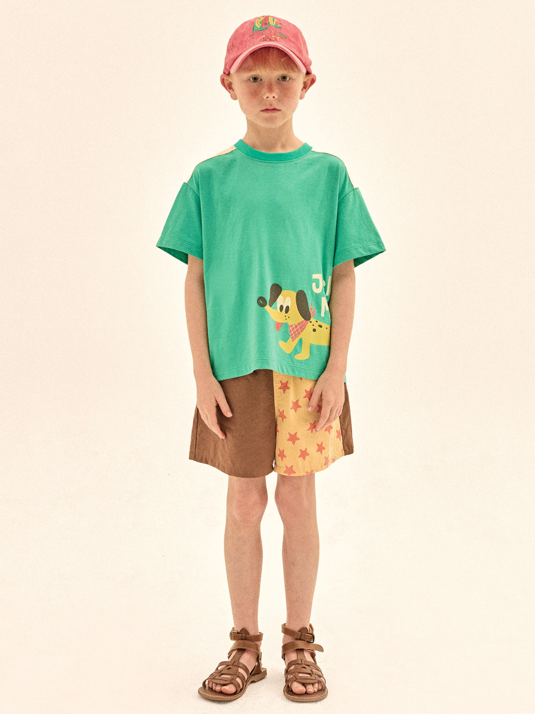Child wearing a green t-shirt with a cartoon character, brown shorts, and a pink cap on a beige background