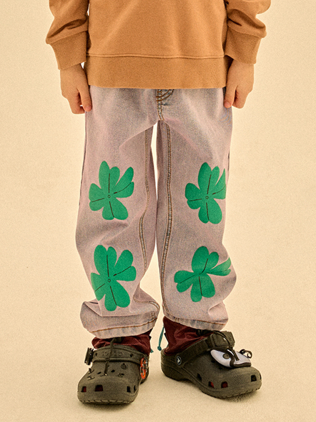 A child in LUCKY CLOVER DENIM PANTS, a brown sweatshirt, and black Crocs with red socks.