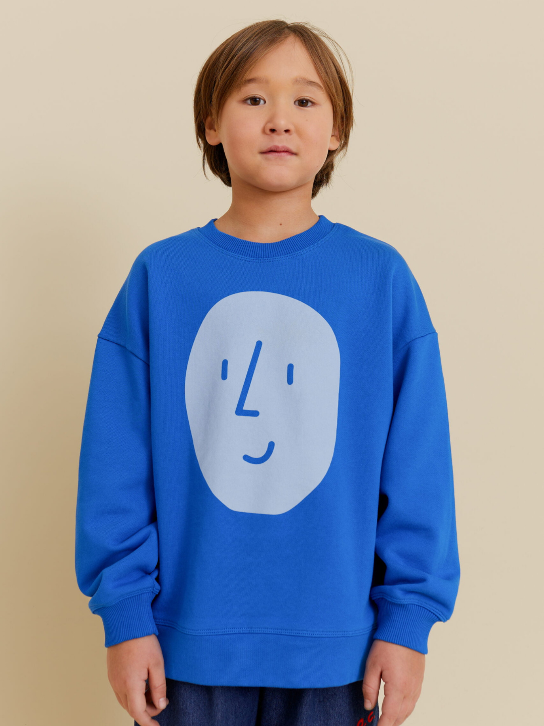 Blue | A child stands against a plain beige background, wearing the POTATO SWEATSHIRT by OXOX CLUB—an oversized, organic cotton sweatshirt featuring a simple cartoon face design on the front.