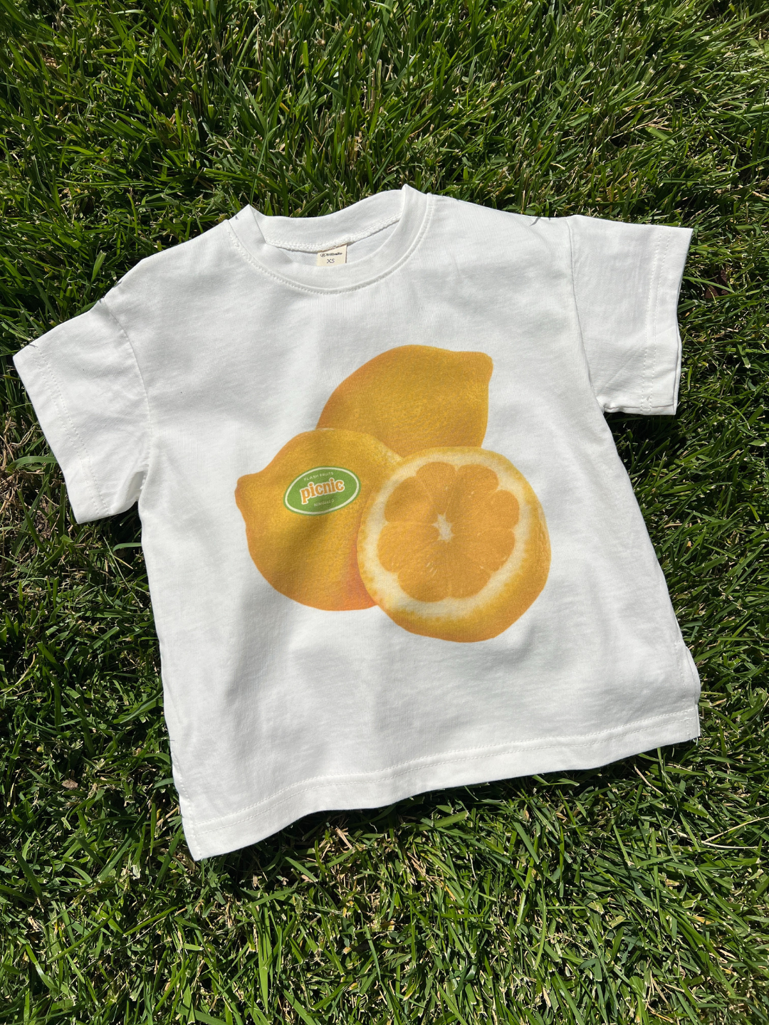 PICNIC TEE: An oversized white cotton t-shirt featuring a screen-printed design of two lemons, one whole and one halved, resting on green grass.