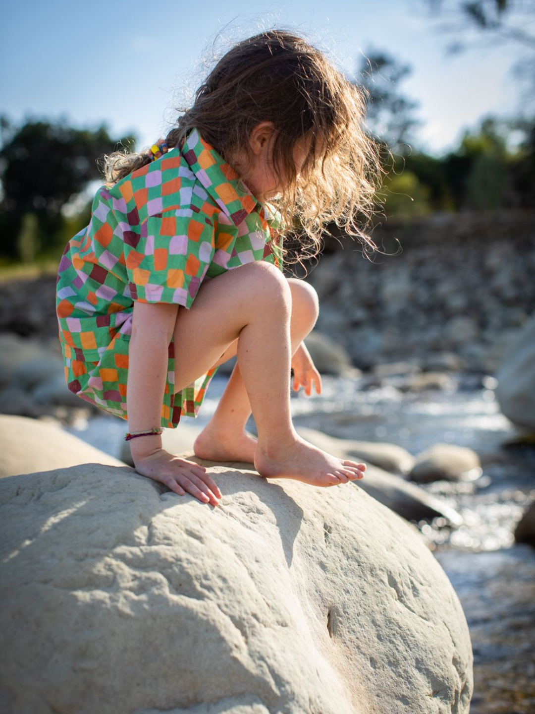 Green | A child on a rock, wearing a kids' shirt and shorts set in a pattern of purple, pink, gold and orange squares against a green background