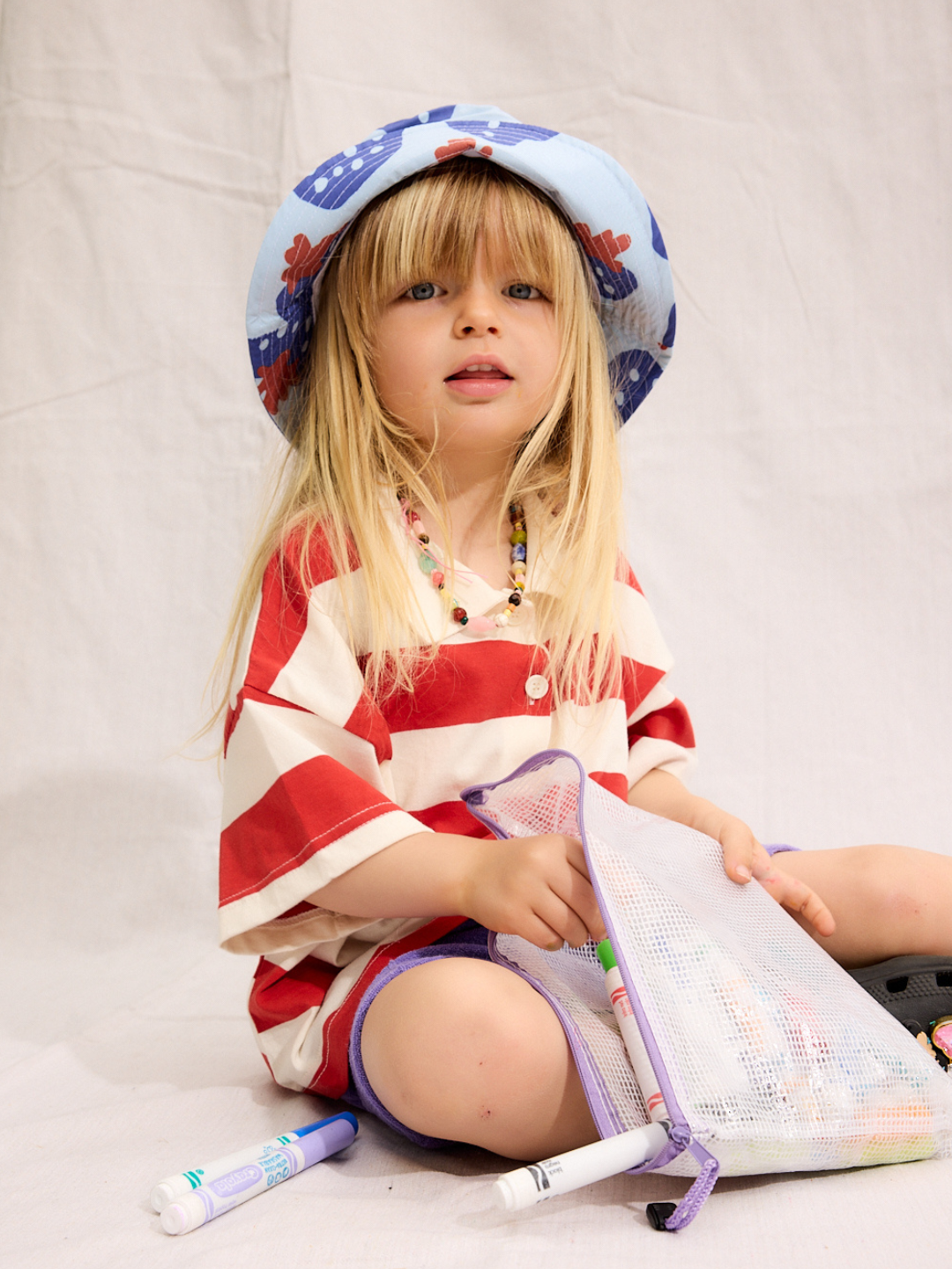 A child is coloring with markers while wearing a Strawberry bucket hat with a red stripe polo shirt
