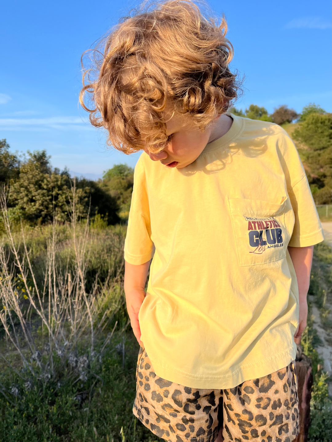 A young child with curly hair stands on a grassy path, looking down while wearing the CLUB POCKET TEE—an oversized yellow top from a Korean kids brand—and leopard print shorts.