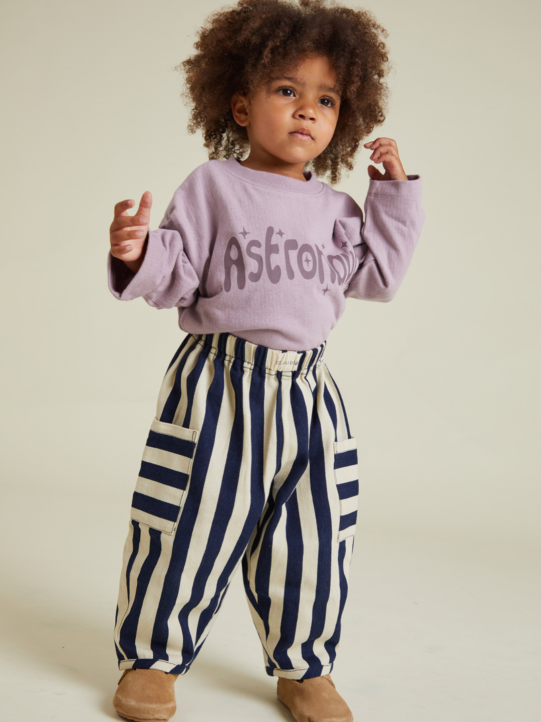 A young child with curly hair wears the EMERSON MIDNIGHT STRIPE TROUSER—loose, striped pants with large pockets—paired with a lavender long-sleeve shirt, ideal for families seeking affordable options and low shipping costs.
