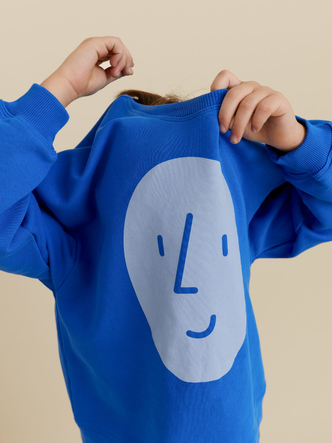 Blue | A child in a roomy POTATO SWEATSHIRT by OXOX CLUB, crafted from organic cotton, lifts the blue top over their head to reveal the playful graphic while hiding their face.