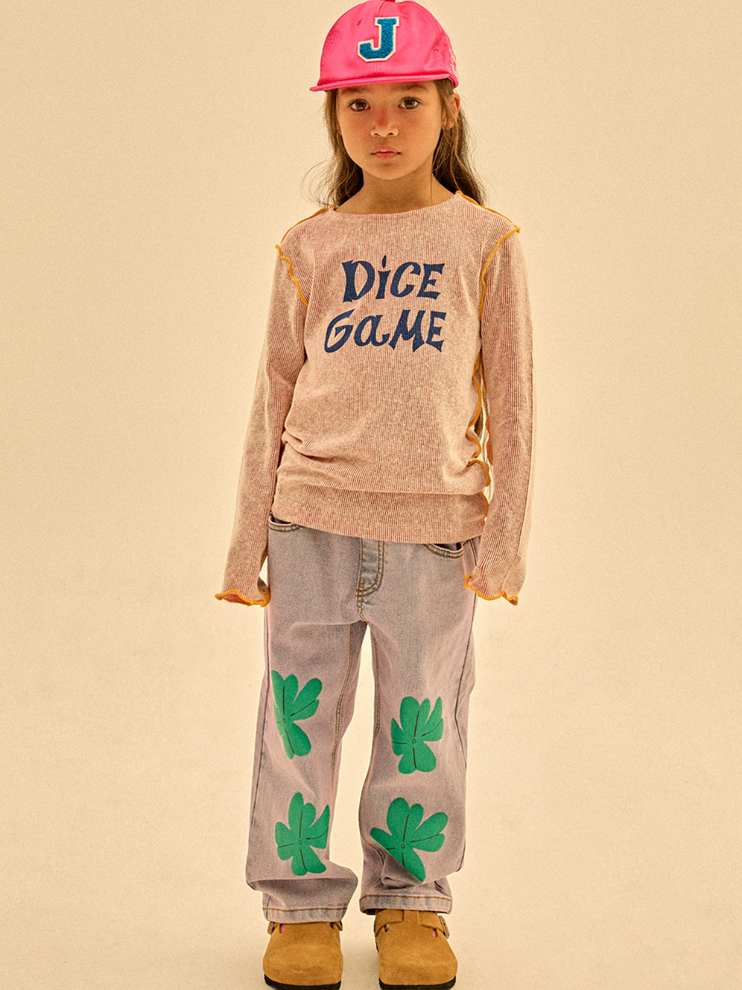 A child in a pink "J" cap, beige "DICE GAME" long-sleeve shirt, LUCKY CLOVER DENIM PANTS with green clover patches, and tan shoes stands against a beige background—proving great style can be affordable.