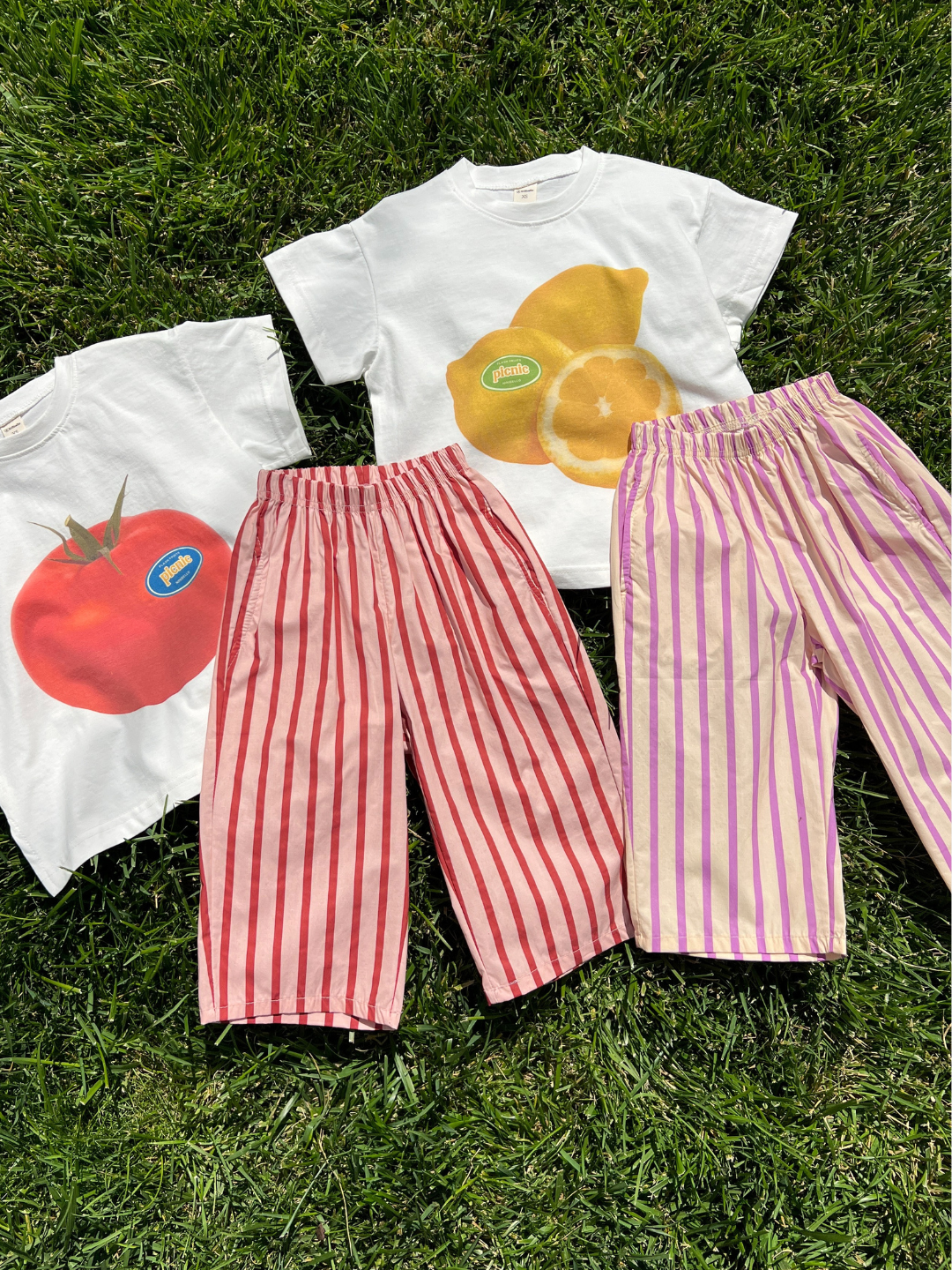 Two oversized PICNIC TEE cotton t-shirts with screen-printed fruit graphics and two pairs of striped pants from a Korean kids brand are displayed on green grass in sunlight.