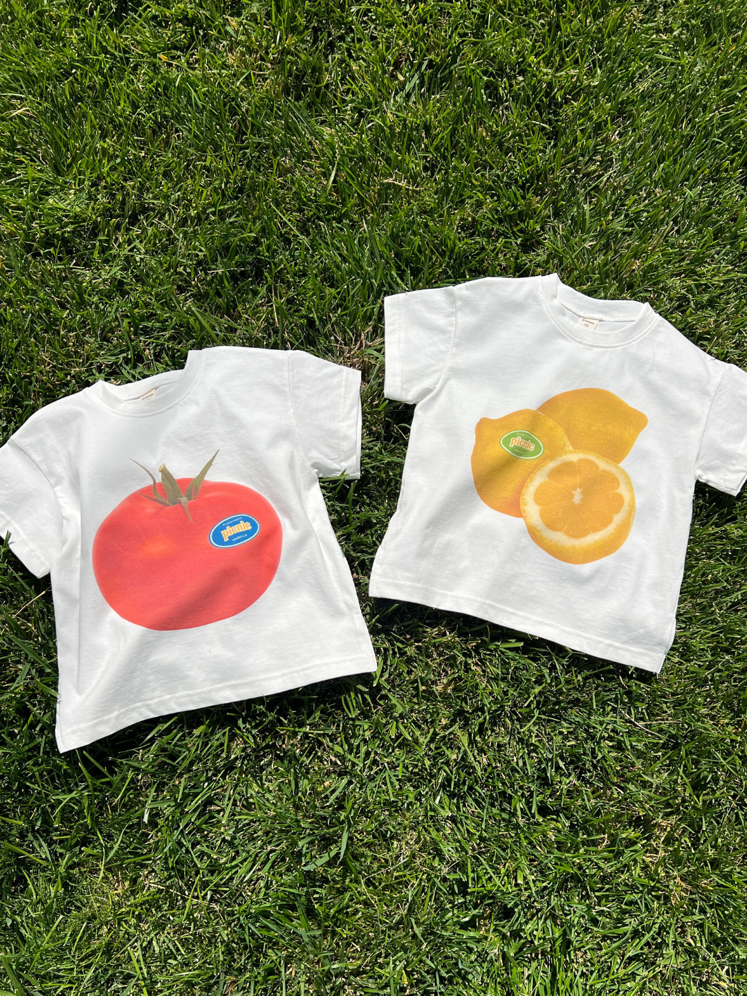The PICNIC TEE, a white oversized cotton t-shirt with a bold tomato print from a Korean kids brand, is paired with the matching t-shirt with a lemon, laid out on green grass.