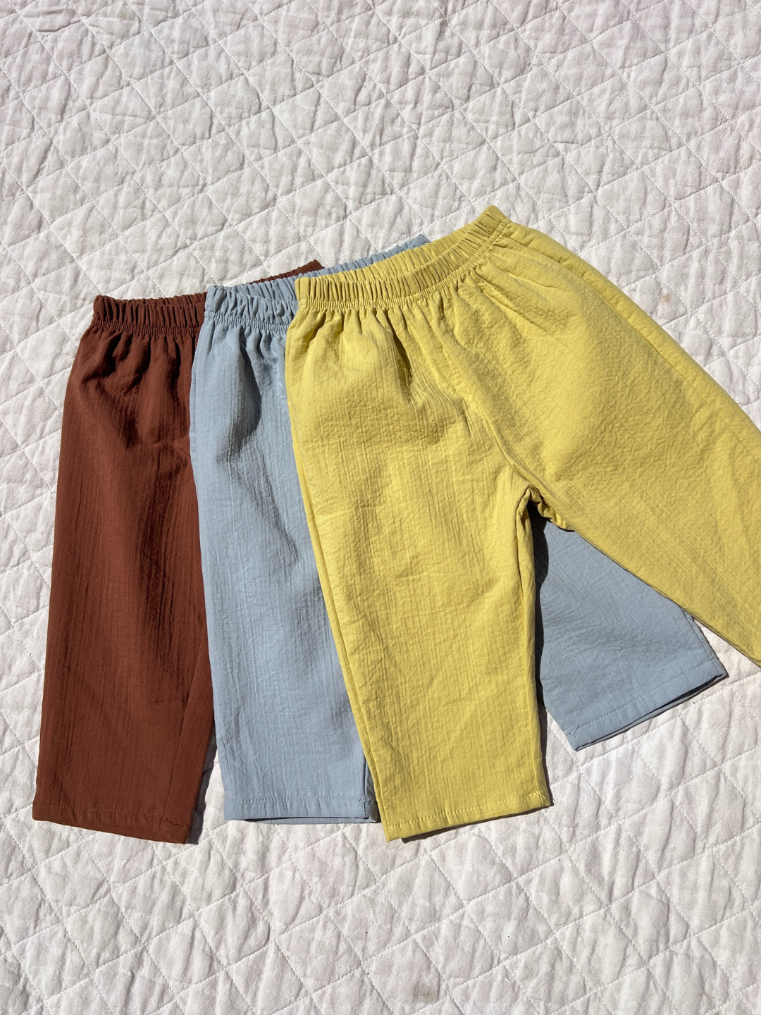 Three pairs of CLOUD COTTON PANT in brown, light blue, and yellow are laid out on a quilted white surface, crafted from breathable woven cotton for all-day comfort.