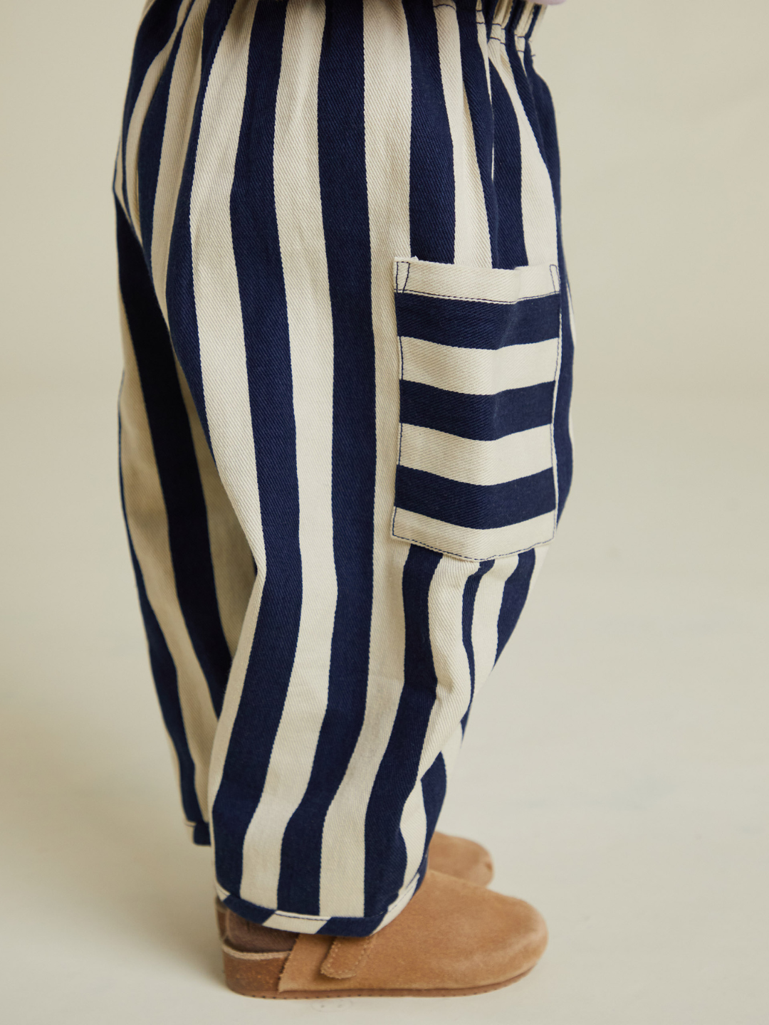 A child wears the EMERSON MIDNIGHT STRIPE TROUSER—beige and navy with a contrasting striped pocket—paired with light brown suede shoes, creating a stylish look for kids, all with free shipping.