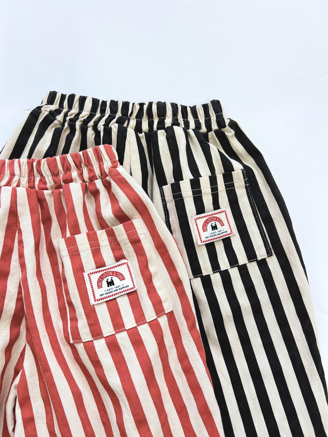 Black/Ecru | Two pairs of LICORICE STRIPE PANT—one red/white, one black/white—in cotton twill with elastic waists and a square patch pocket featuring an elephant logo framed in red.