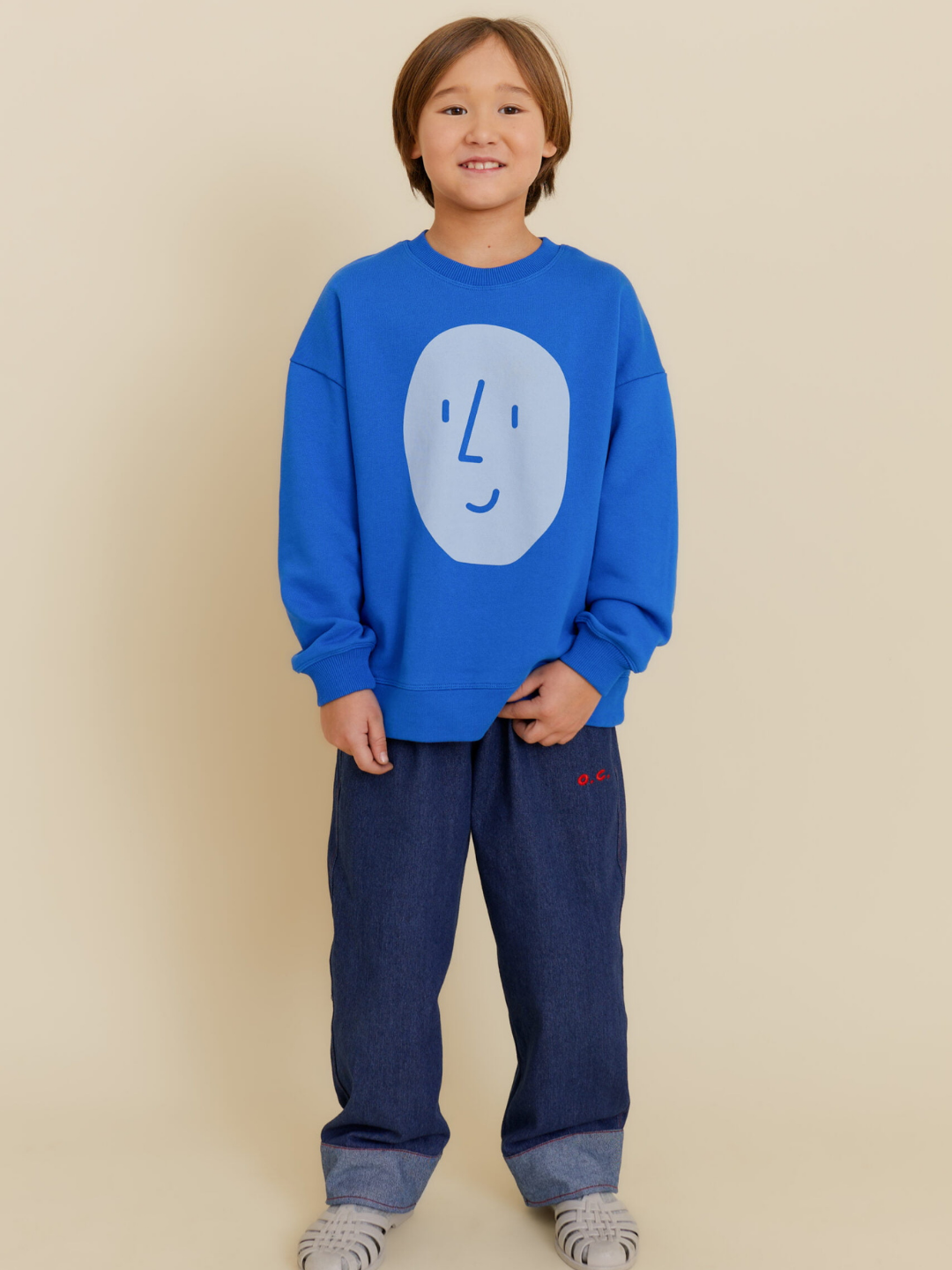 Blue | A child smiles at the camera against a beige background, wearing the POTATO SWEATSHIRT by OXOX CLUB—an oversized, organic cotton top with a simple face graphic—paired with loose blue jeans.