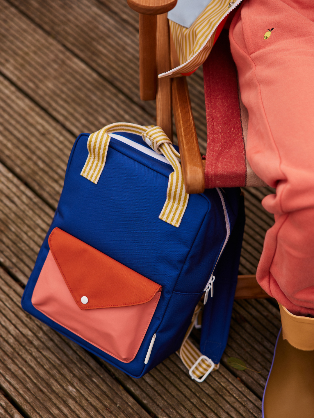The MEDIUM ENVELOPE BACKPACK, made from recycled materials with blue fabric and orange-pink accents, sits on a wooden deck beside a person in pink pants and tan boots on a wooden chair.