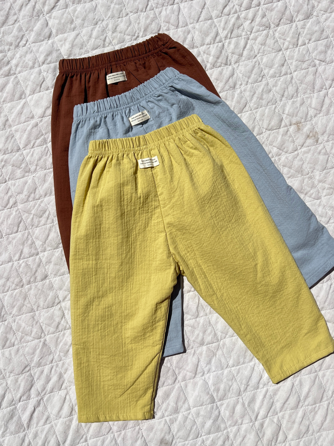 Three pairs of CLOUD COTTON PANT in yellow, light blue, and brown are displayed on a quilted white surface. Made from breathable woven cotton, these relaxed fit children’s pants provide all-day comfort.