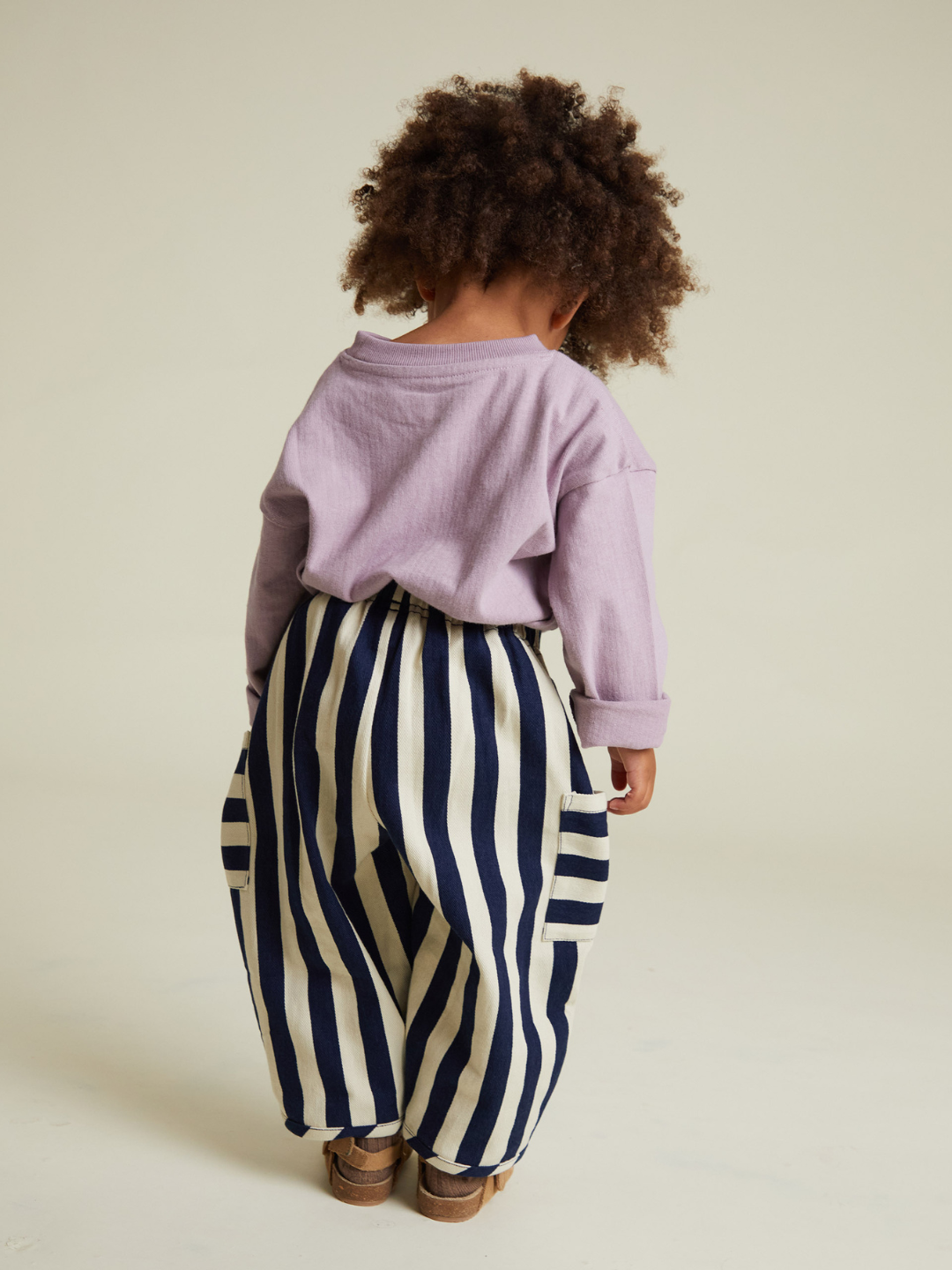 A young child with curly hair faces away, wearing a lavender long-sleeve top and EMERSON MIDNIGHT STRIPE TROUSER—loose, high-waisted pants with navy and white stripes for stylish comfort without extra shipping costs.