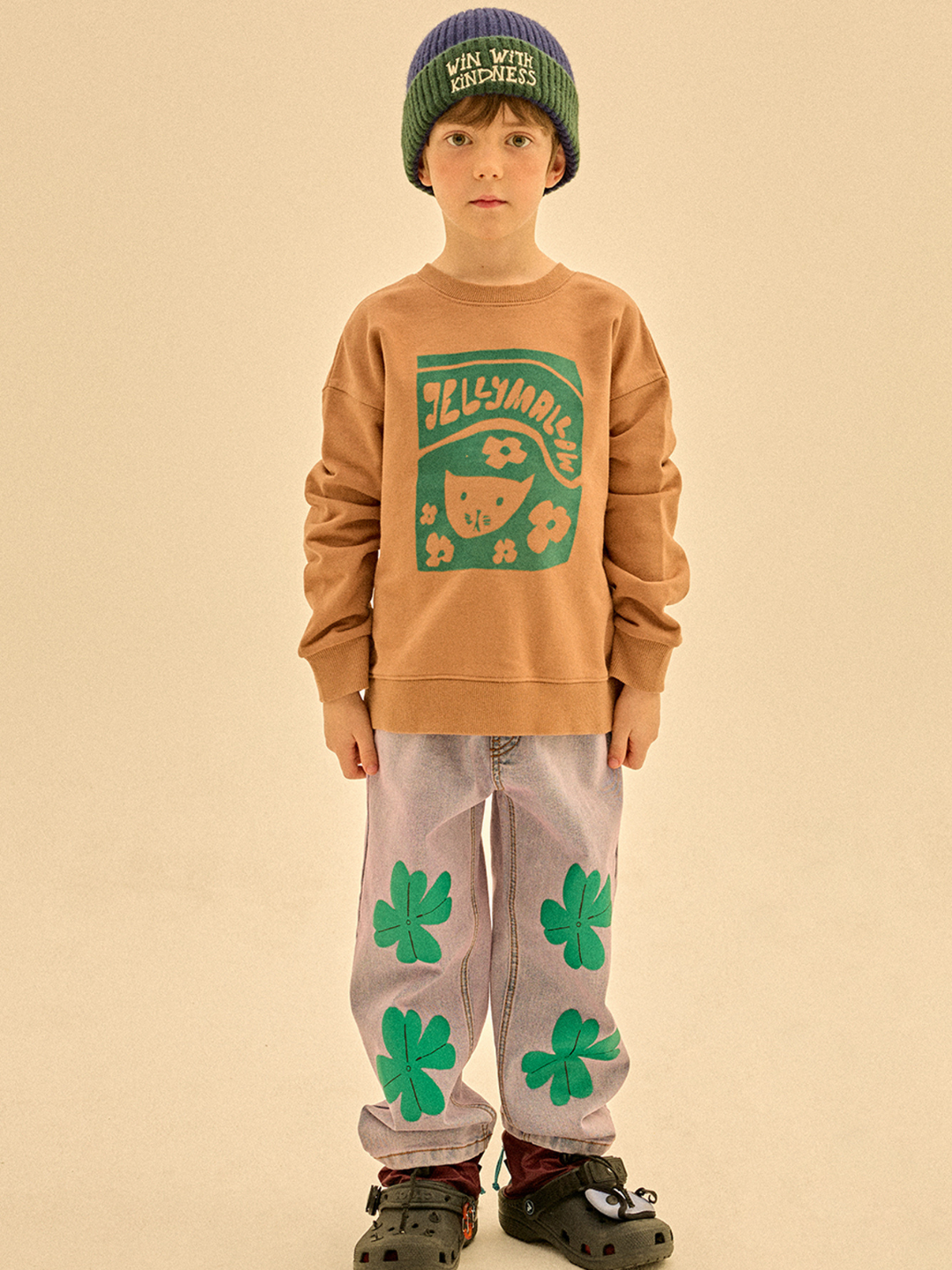 A child faces forward in LUCKY CLOVER DENIM PANTS—light jeans with green clover prints—paired with a green beanie, tan graphic sweater, and dark slip-on shoes, all set against a simple beige background.