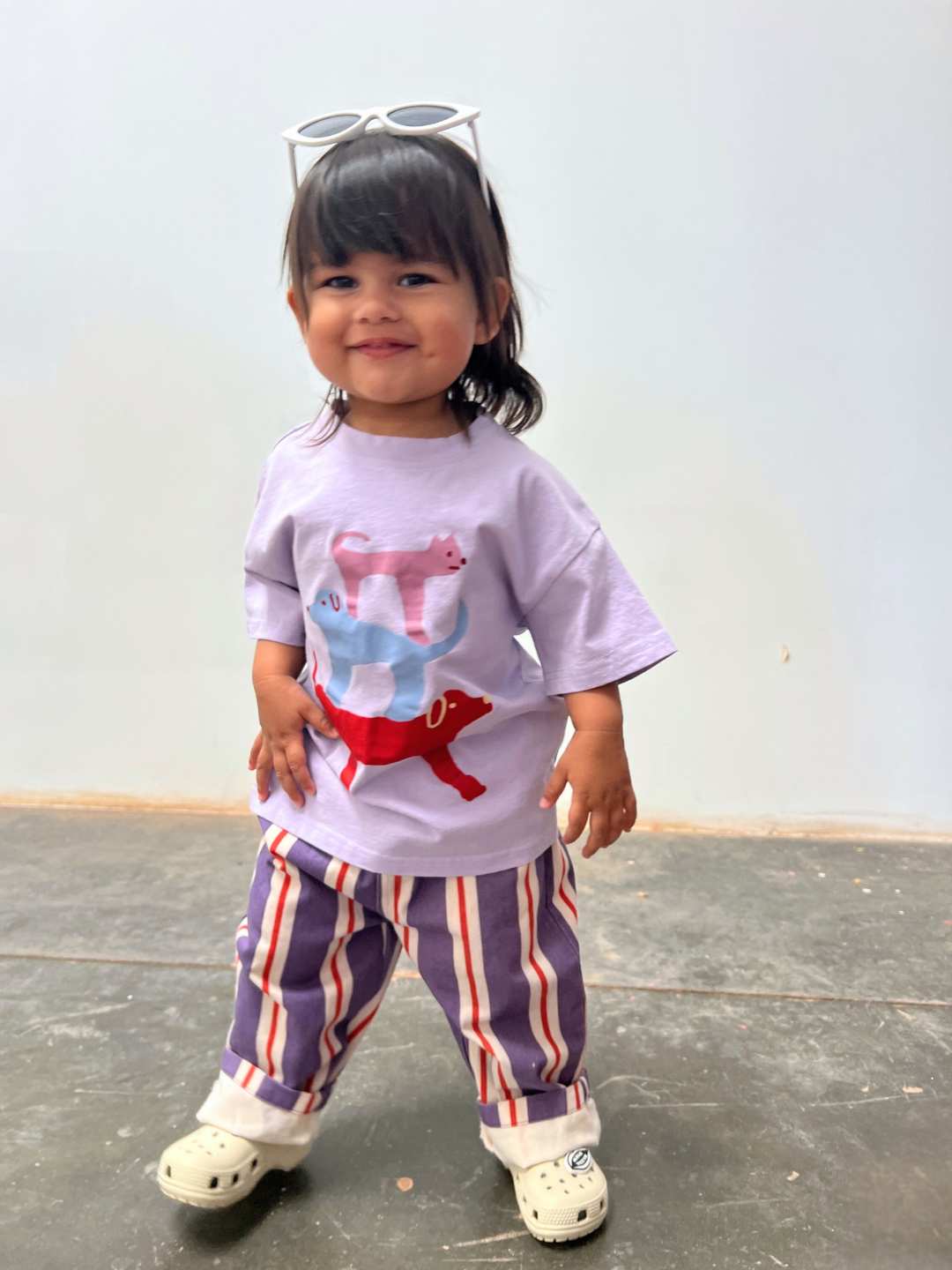 A young child smiles indoors at the camera in a pastel purple shirt with colorful animal graphics, STRIPE TWILL PANT, white shoes, and sunglasses on their head.