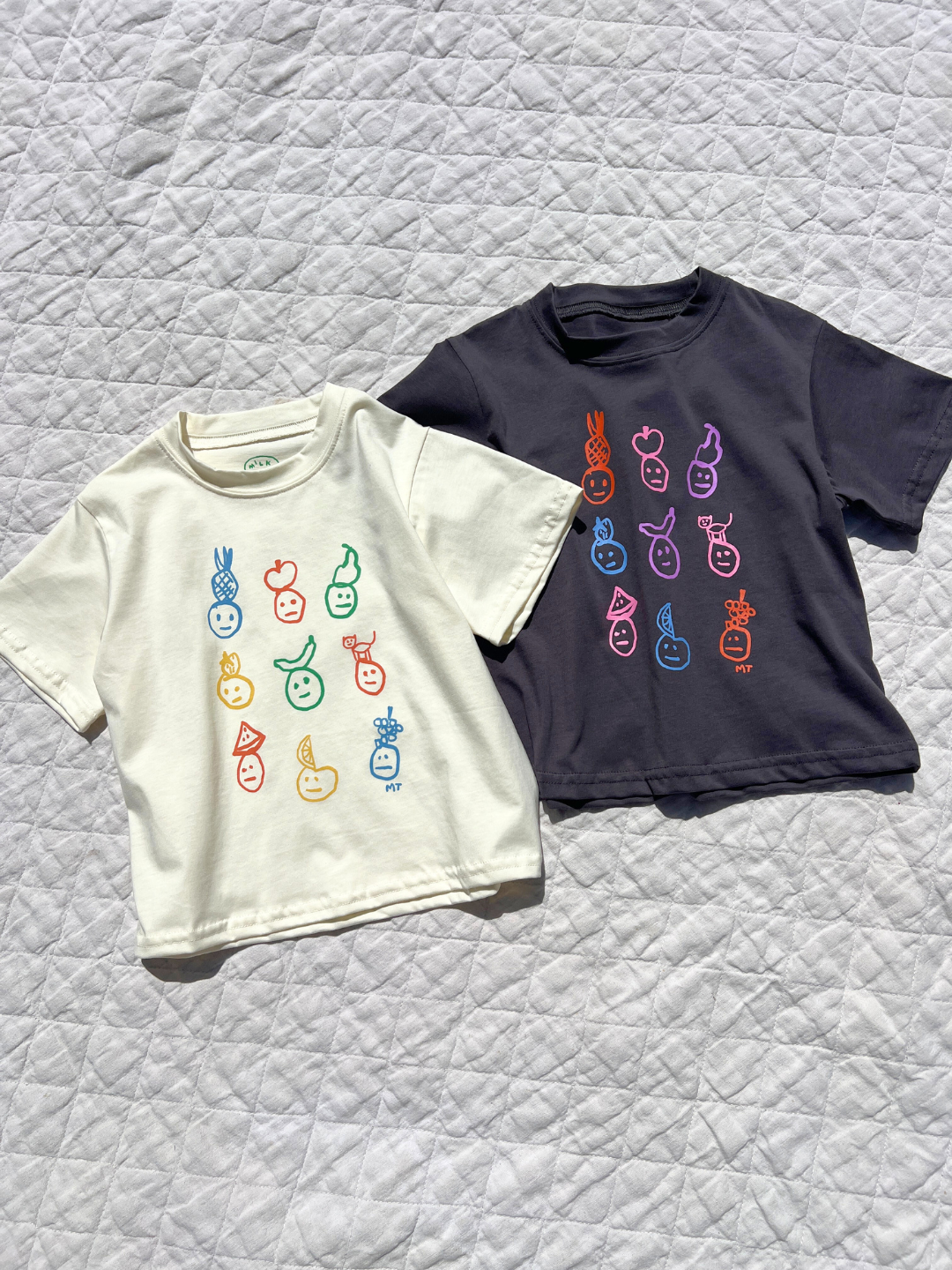 Cream and charcoal color ways of fruit dace tees laid flat on the blanket