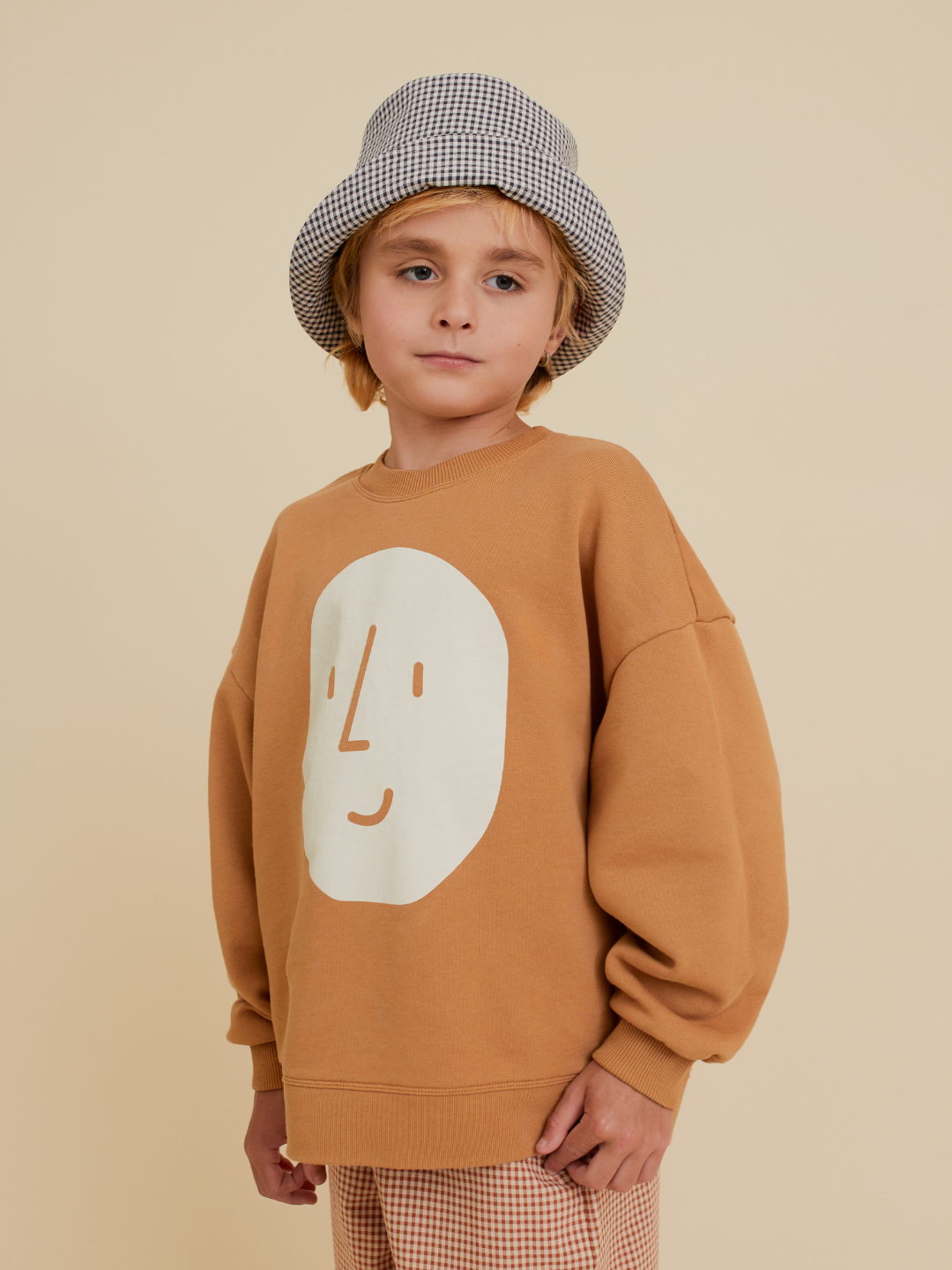 Brown | A young child in an OXOX CLUB POTATO SWEATSHIRT made from organic cotton, paired with a checkered bucket hat and plaid pants, stands against a beige background.