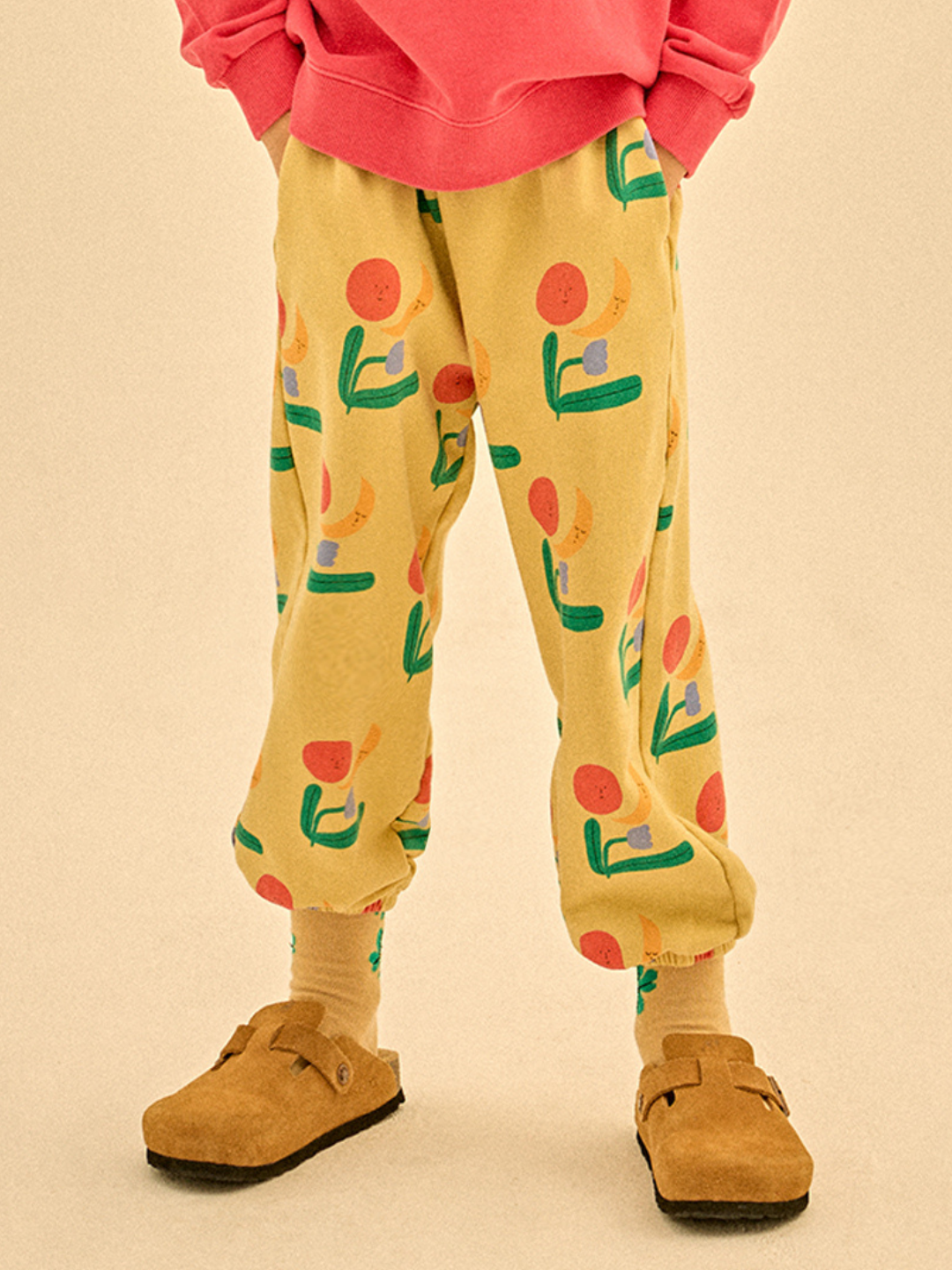 Wearing COLORED NATURE ALLADIN PANTS—yellow with colorful abstract designs—a model in a pink sweatshirt, beige socks, and tan clogs stands against a plain background