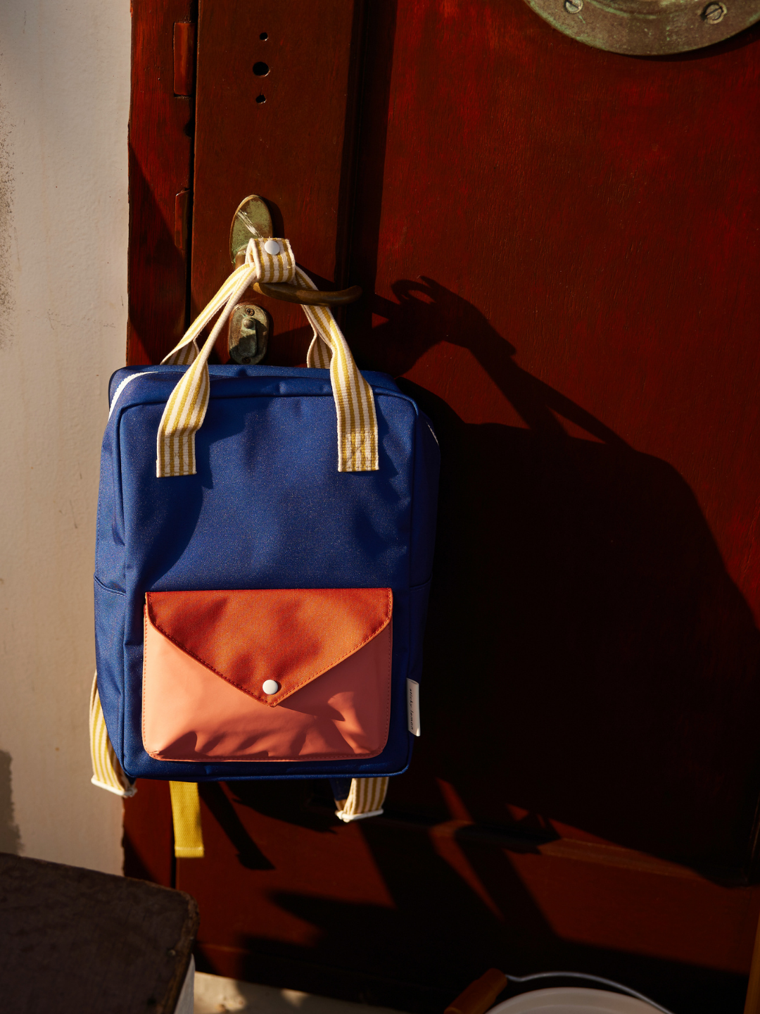 Ned Blue | The MEDIUM ENVELOPE BACKPACK, crafted from recycled materials with beige straps and an orange front pocket, hangs on a door handle in bright sunlight.