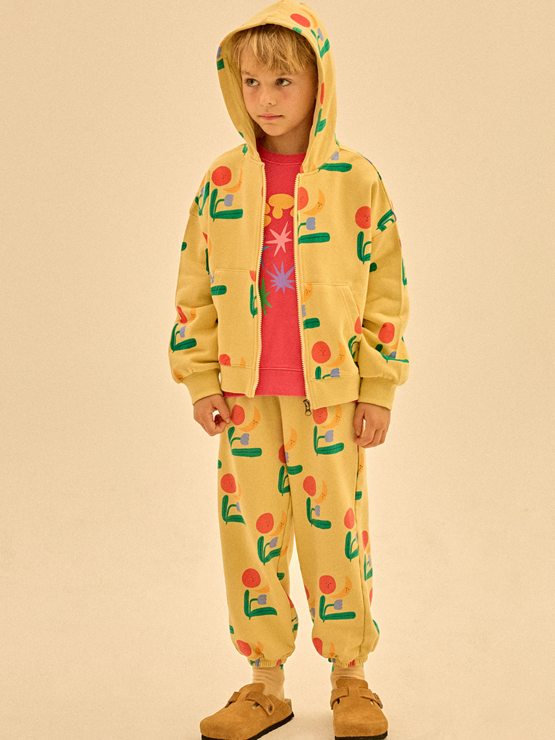 A child models the COLORED NATURE ALLADIN PANTS—yellow with vibrant abstract prints—paired with a pink tee and brown slip-ons, effortlessly displaying playful style