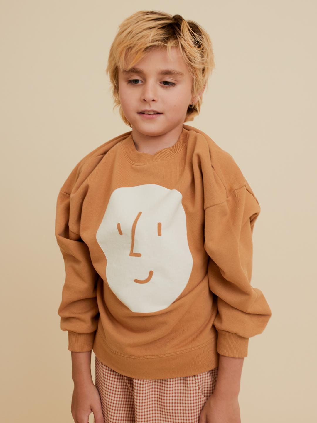 Brown | A child with blonde hair wears the oversized, organic cotton POTATO SWEATSHIRT by OXOX CLUB, featuring a simple abstract face, paired with checkered pants and standing against a beige background.