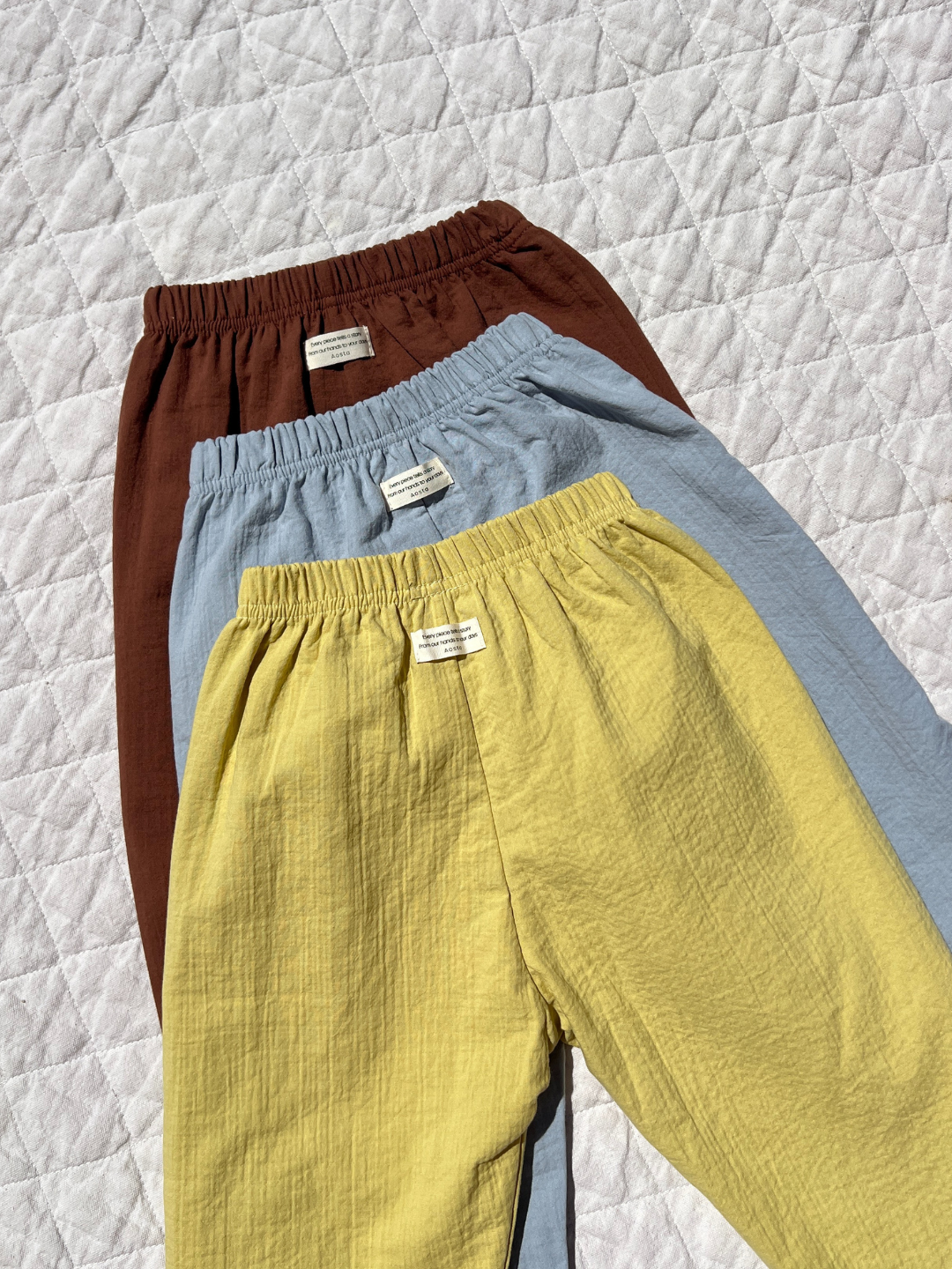 Three pairs of CLOUD COTTON PANT in brown, light blue, and yellow are arranged on a quilted white surface. Made from breathable double-layer woven cotton, they offer effortless style and comfort.