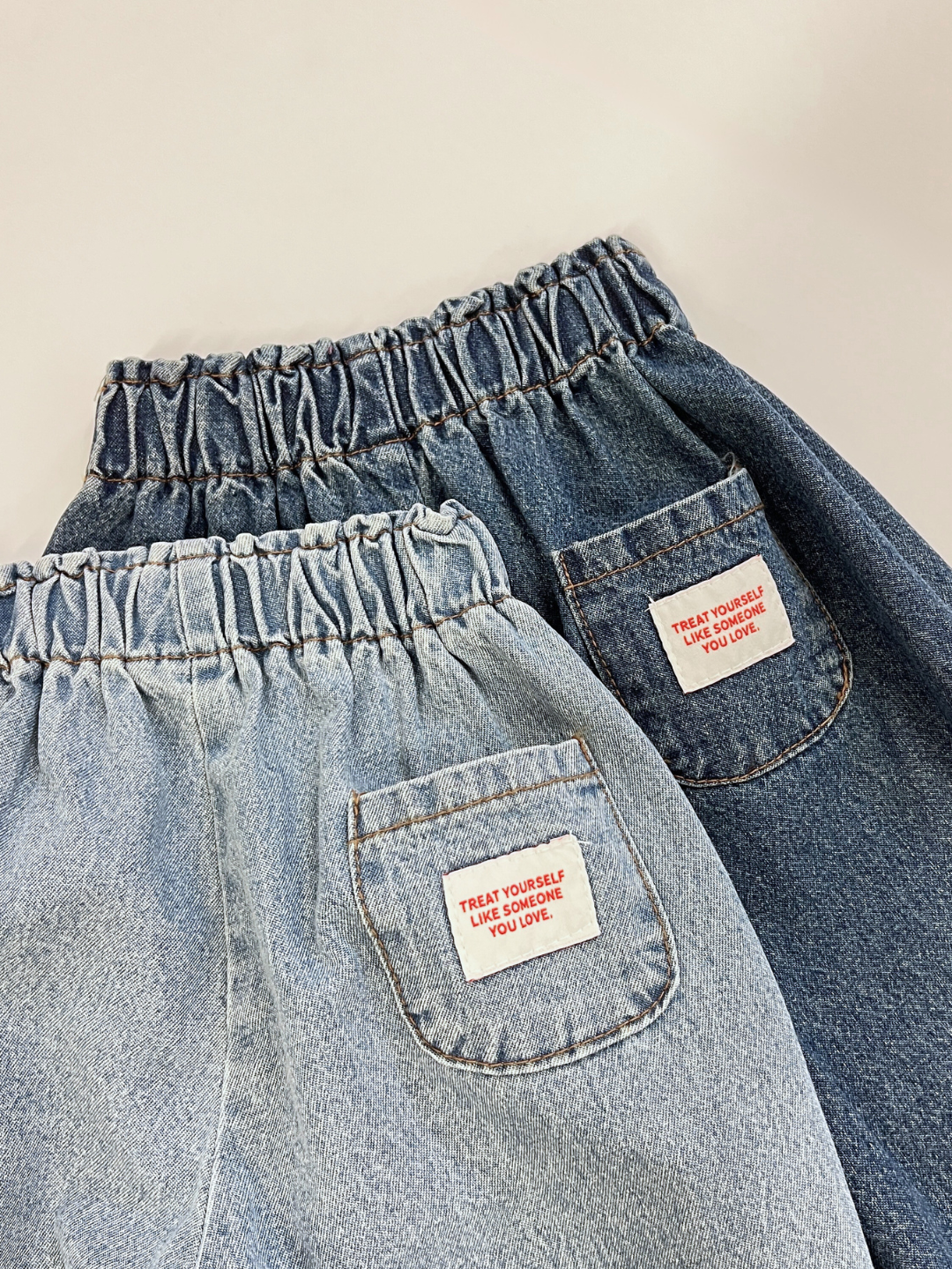 Dark Wash | BABY JEANS: Set of 2 Korean cotton denim baby jeans—one light blue, one dark blue—both with elastic waistbands and back pocket labels that say, “Treat yourself like someone you love.”.