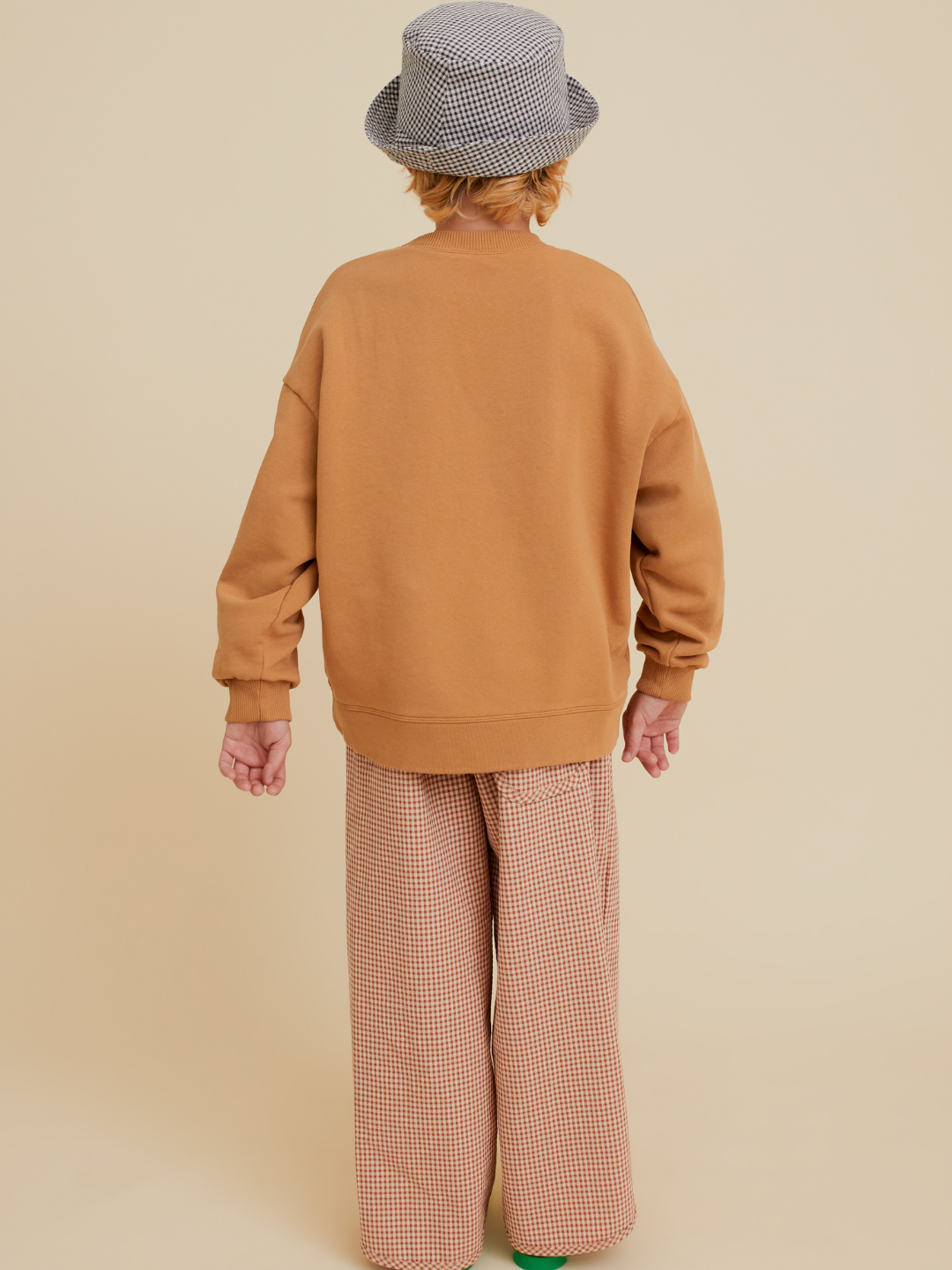 Brown | Child with back to camera wears an OXOX CLUB POTATO SWEATSHIRT made of organic cotton, paired with patterned wide-leg pants and a textured gray hat, set against a beige background.