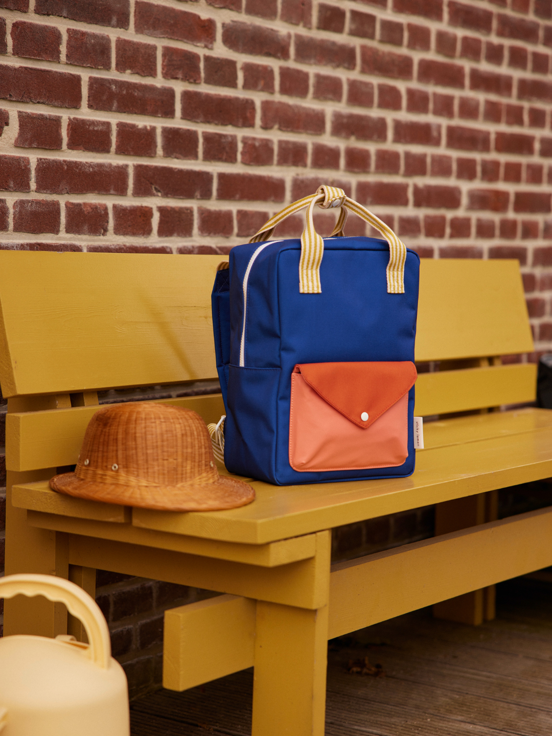 Ned Blue | A MEDIUM ENVELOPE BACKPACK in blue, made from recycled materials with a peach front pocket, and a brown hat sit on a yellow wooden bench against a brick wall.