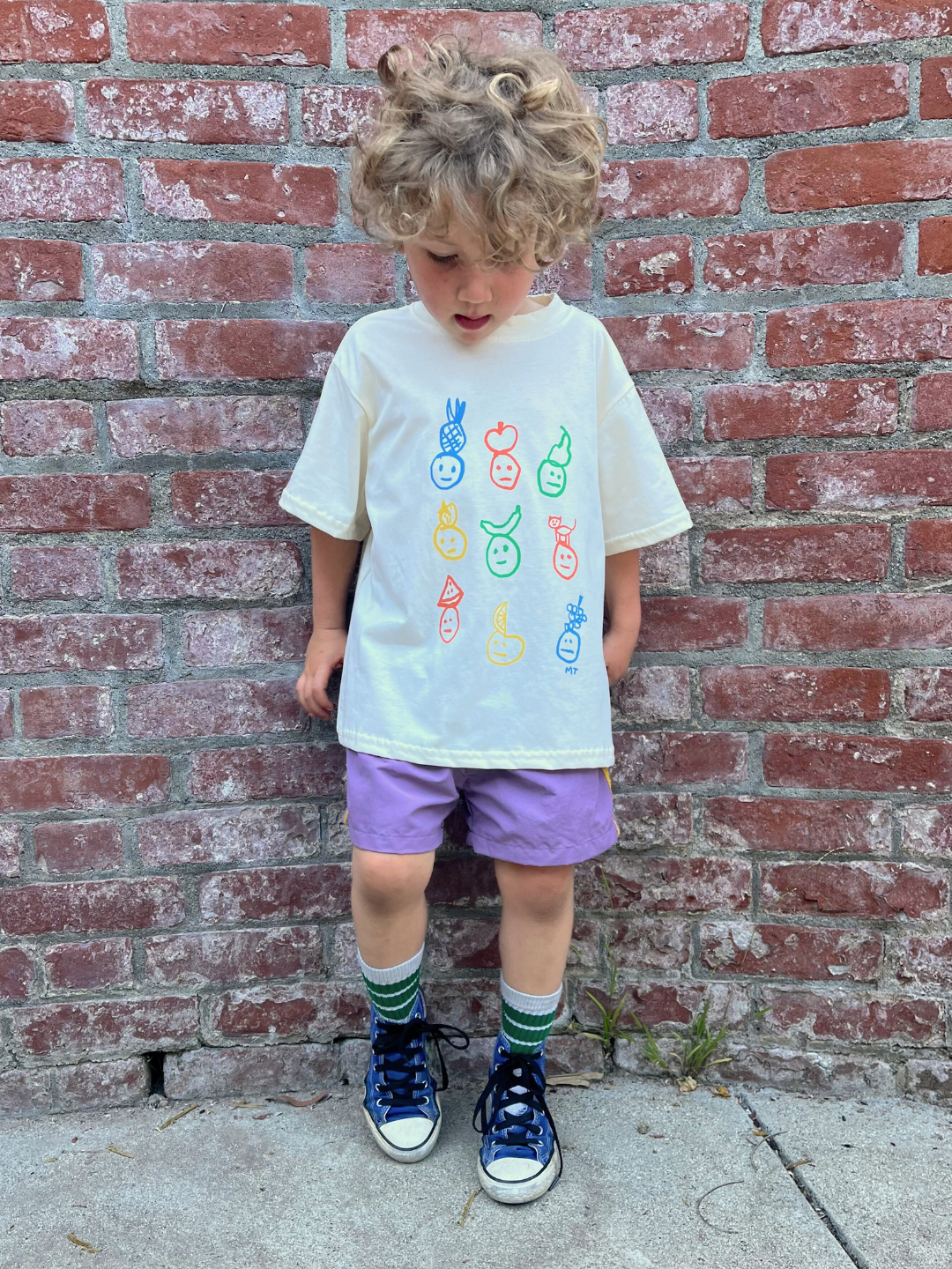A child Is wearing an ivory colorway of the fruit face tee with purple shorts.