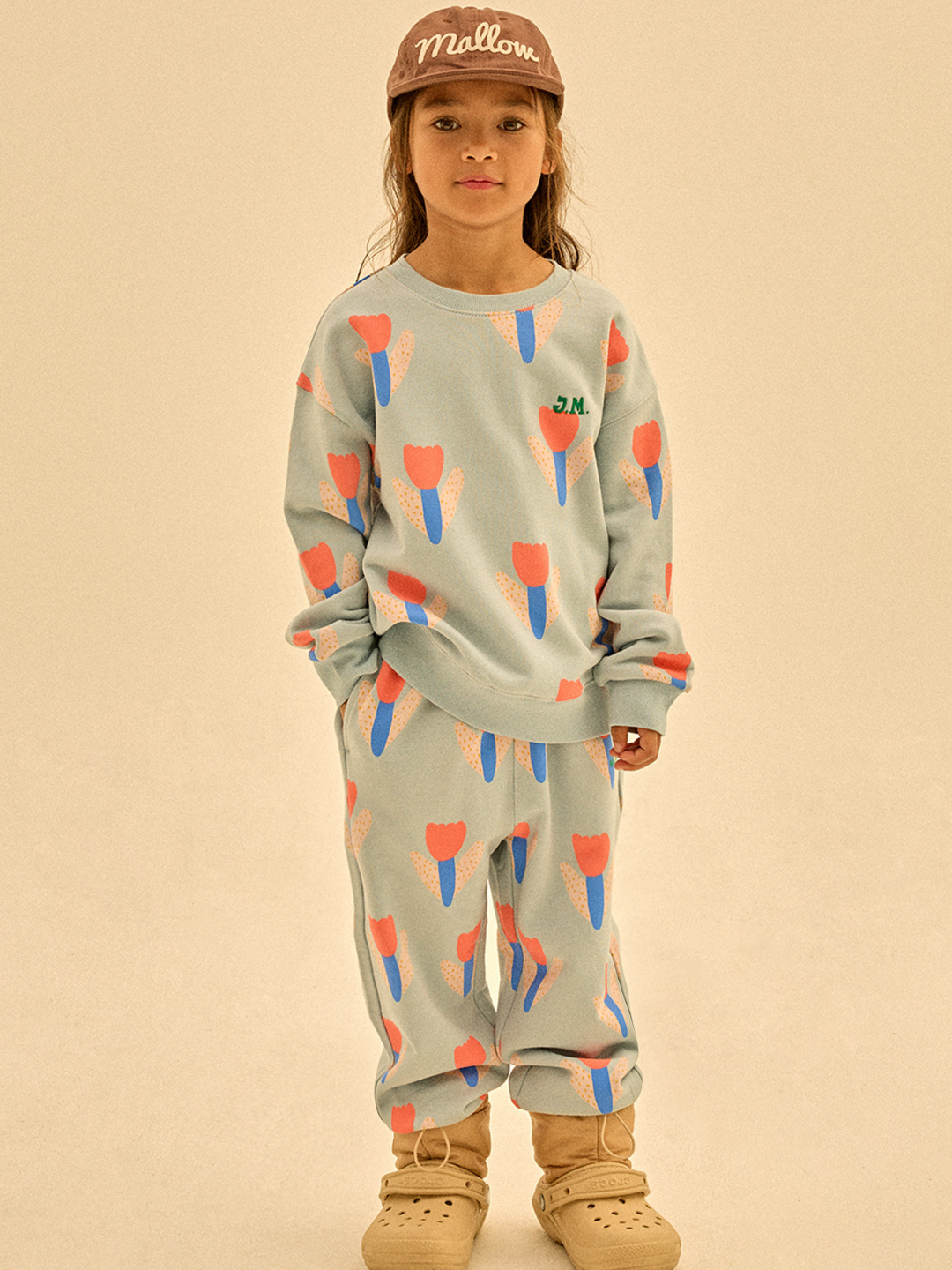 A child wears TULIP SWEATPANTS with pink and blue tulip patterns, tan slippers, and a brown “Mallow” cap, standing against a plain background with a curious look.