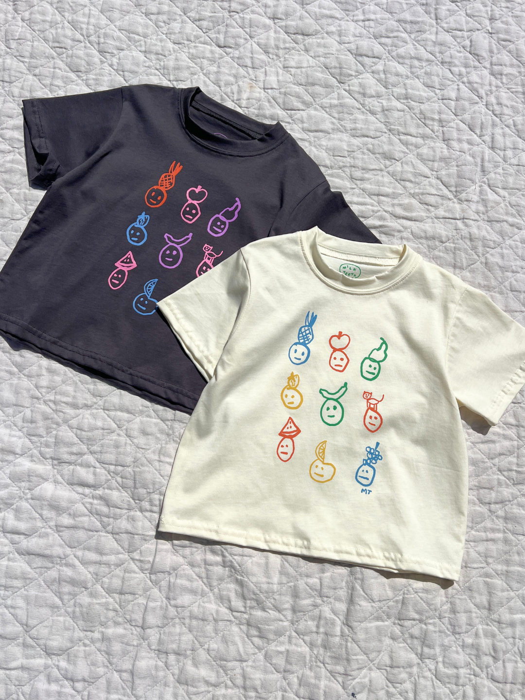 Both colorwasy of the fruit face tee are laid flat on a quilt