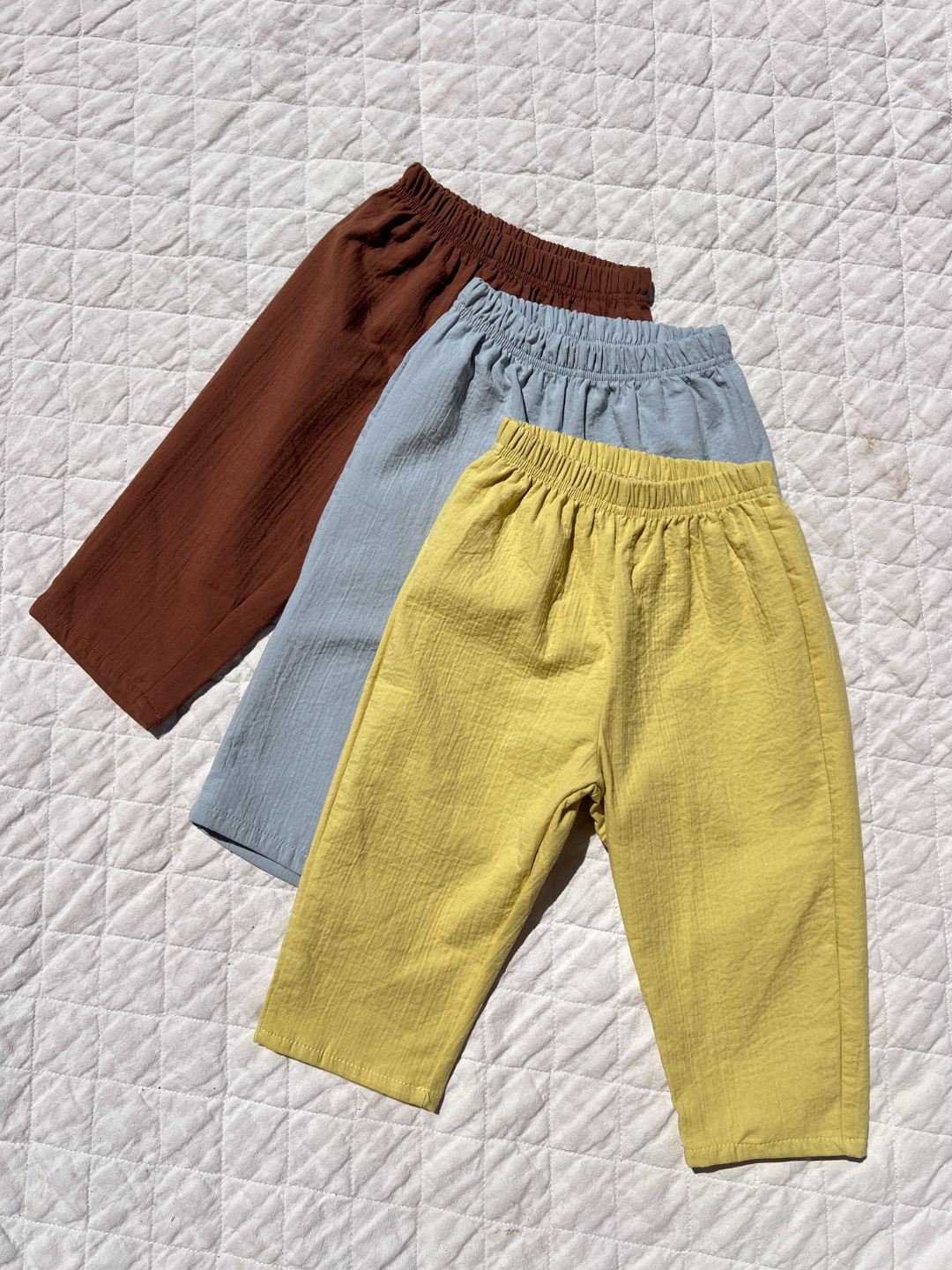 Three CLOUD COTTON PANTs in brown, light blue, and yellow are displayed on a white quilted surface, each crafted from breathable woven cotton for all-day comfort.