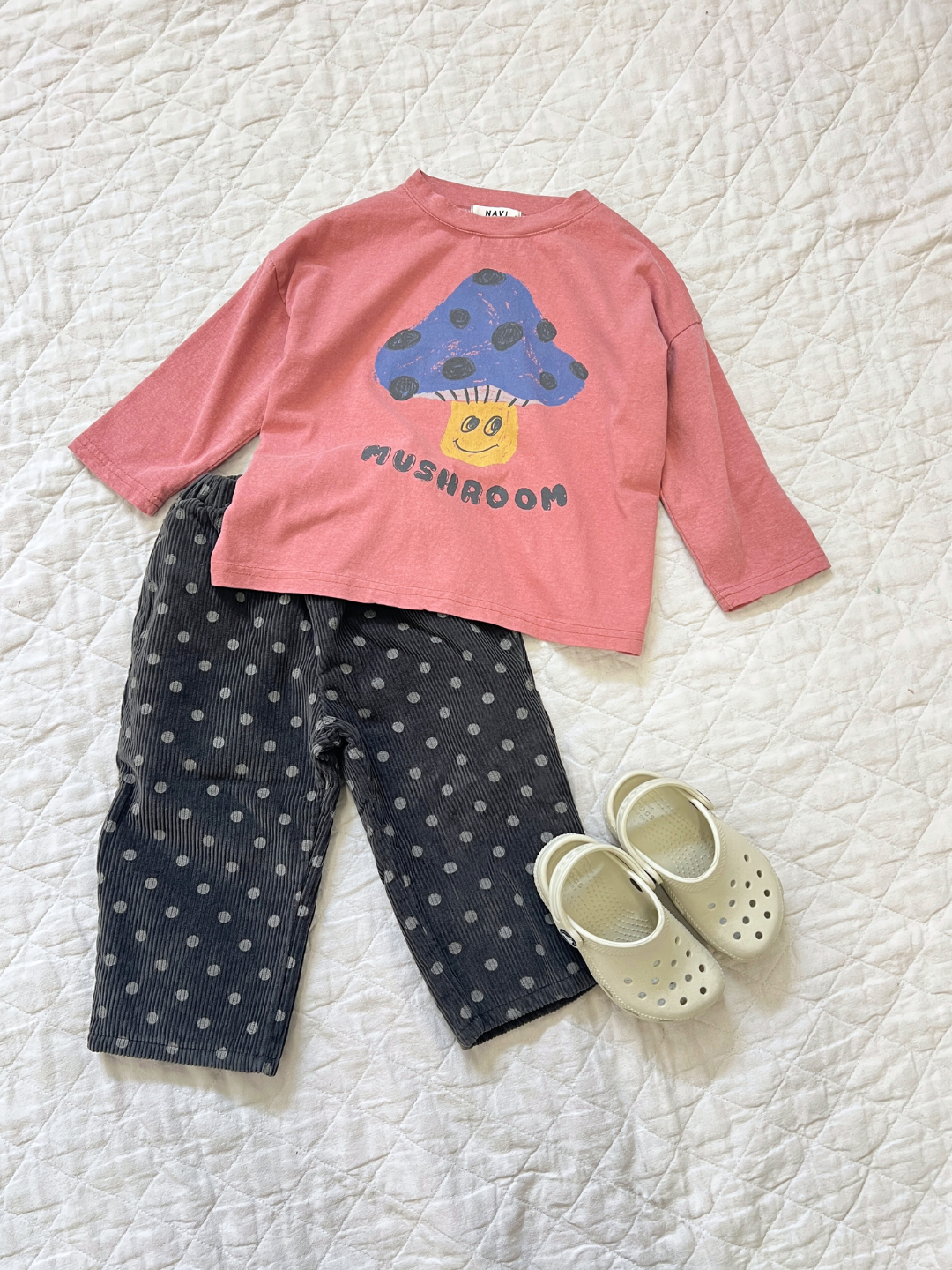 Faded Red | | Children's outfit with a MUSHROOM LONG SLEEVE TEE in pink, adorned with a blue mushroom graphic, paired with gray polka dot pants and white slip-on shoes, all set on a white quilted background.