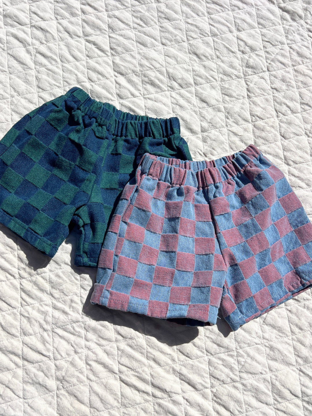 Pink | Two pairs of TWO TONE DENIM CHECK SHORTS—a Korean kids brand style in green/navy and pink/light blue—are displayed on a quilted white surface.