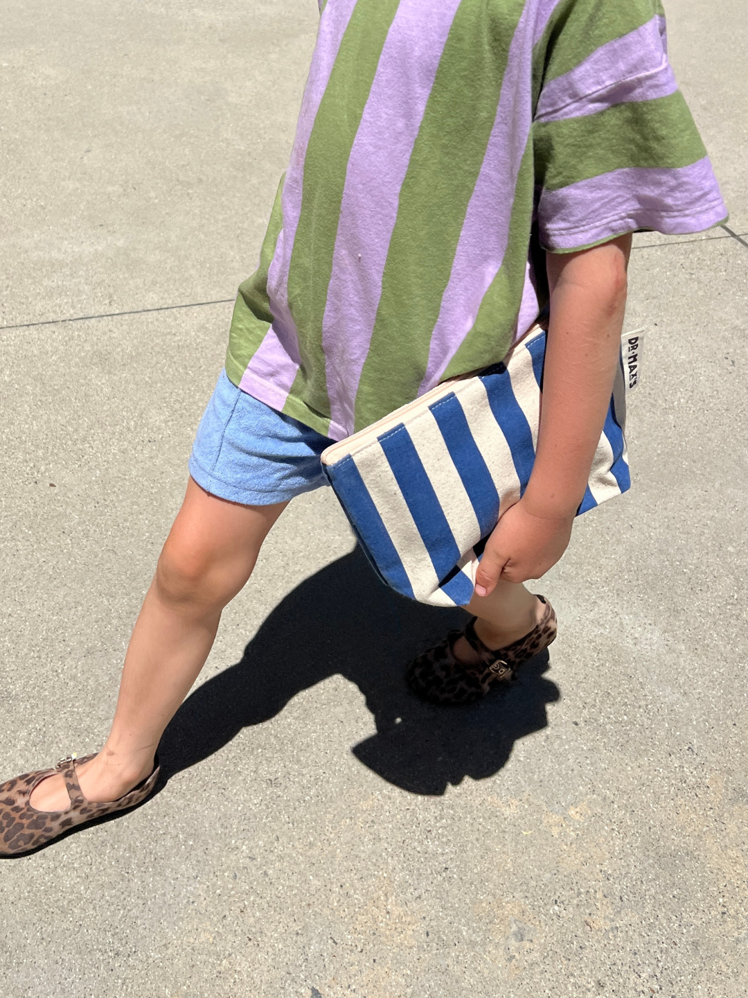 Child wearing a green and purple striped shirt with blue shorts, holding a blue and white striped bag.