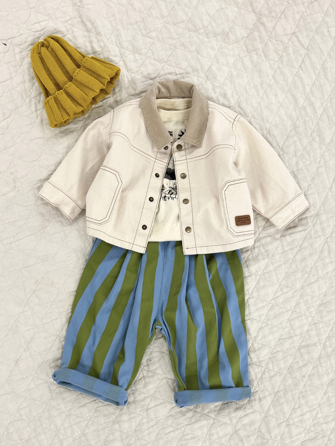 Green | Front view of kids baggy pants in blue with wide green vertical stripes. Shown flat on a beige quilt, paired with an ecru denim jacket, white t-shirt and a yellow beanie.