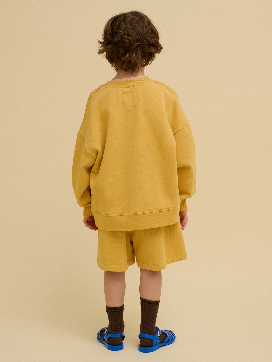 A child stands facing away, wearing a loose yellow FACE SWEATSHIRT, matching shorts, brown socks, and blue sandals against a beige background.