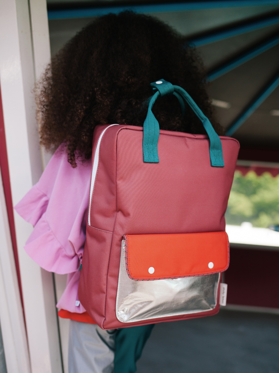 A person with curly hair in a pink top carries the LARGE BETTER TOGETHER ENVELOPE BACKPACK, a large red and orange recycled PET bag with green adjustable shoulder handles, near a striped-ceiling structure.