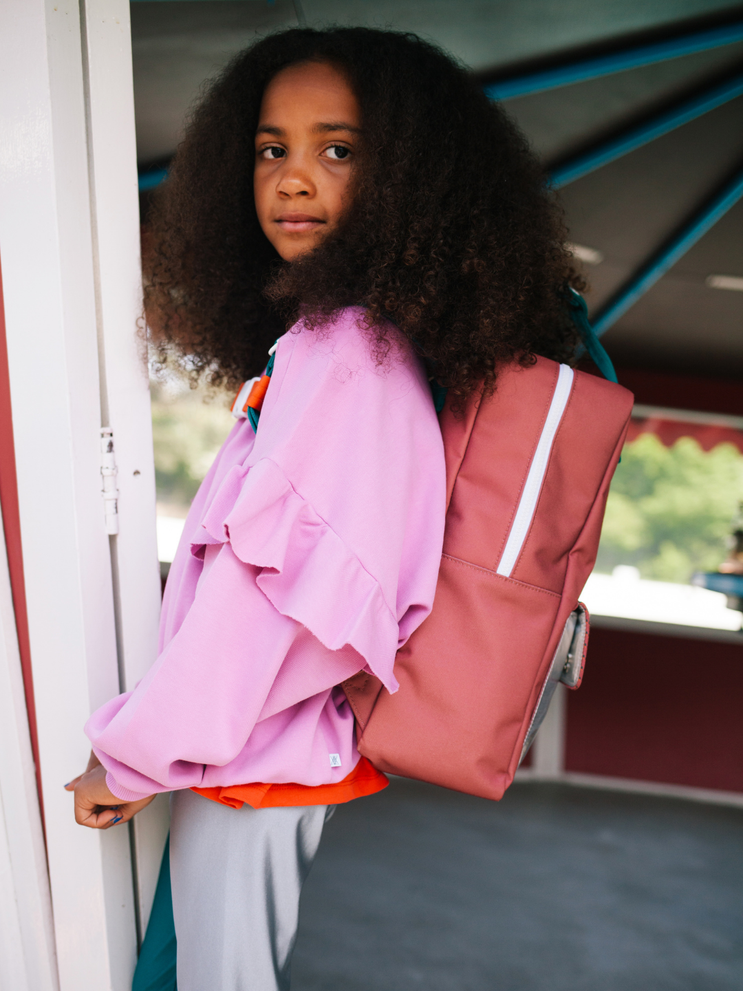 A girl with curly hair, wearing a pink ruffled sweatshirt and the LARGE BETTER TOGETHER ENVELOPE BACKPACK with adjustable shoulder handles, stands near a doorway looking slightly toward the camera.