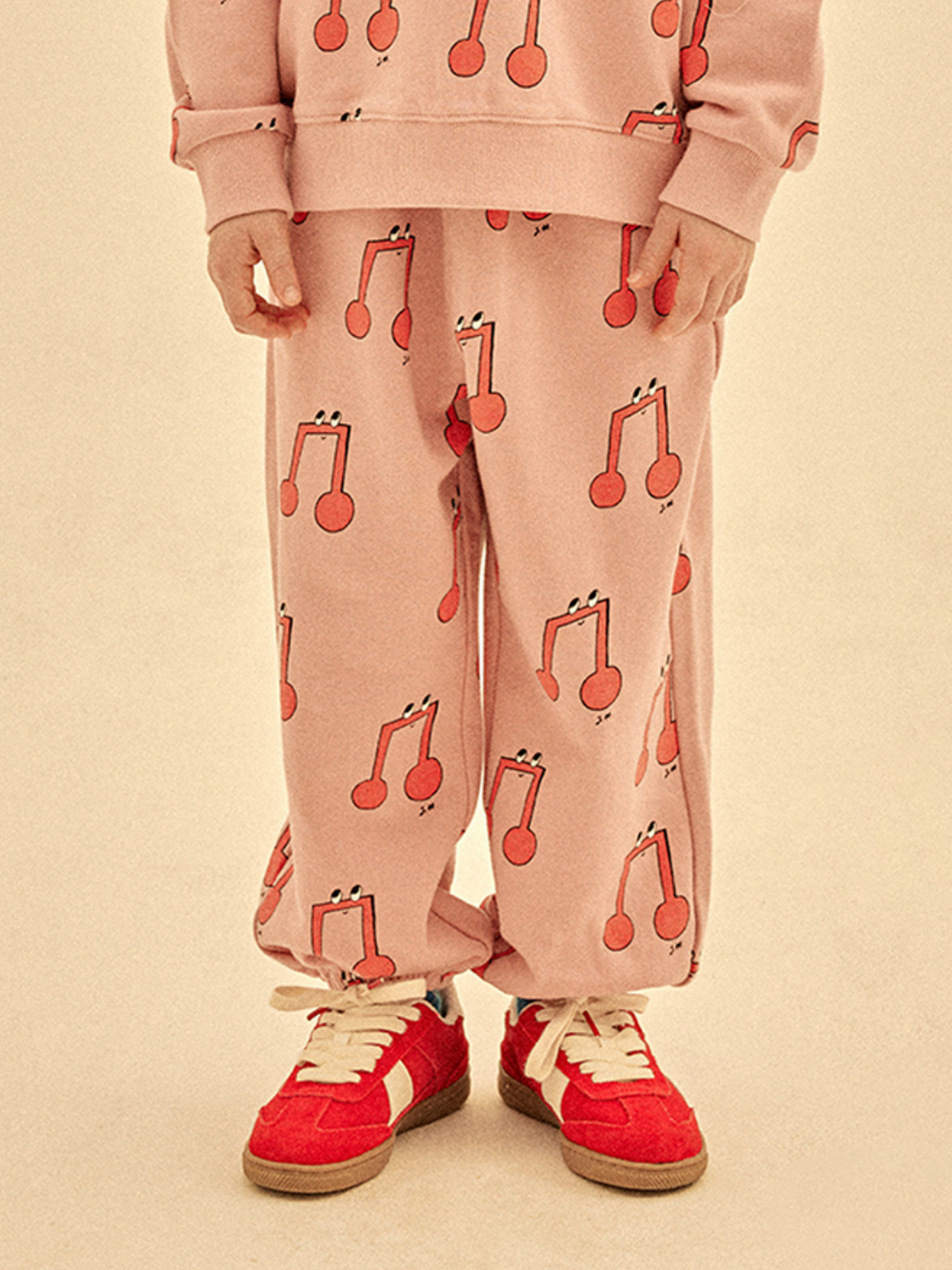 Wearing MELODY ALADDIN PANTS with red musical note print, a matching pink sweatshirt, and red sneakers, the person stands against a beige background
