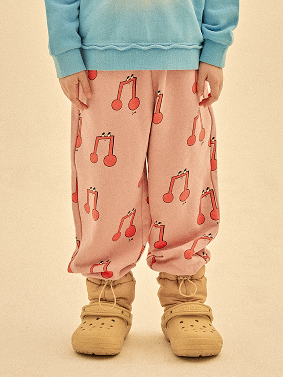 A person wears a blue sweatshirt, beige slip-on shoes, and MELODY ALADDIN PANTS—pink pants featuring red musical note patterns—while standing on a plain background